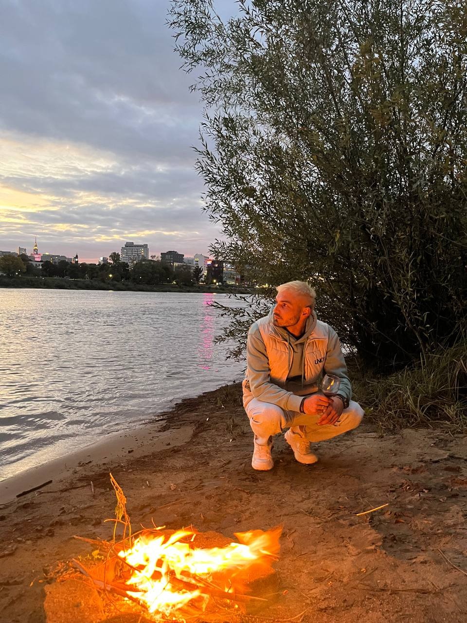 Mężczyzna w jasnej odzieży kuca na piaszczystym brzegu rzeki obok ogniska, trzymając kieliszek wina, w tle widoczne budynki miejskie pod wieczornym niebem.