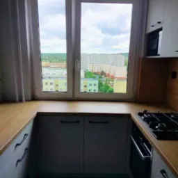 Nowoczesna kuchnia z oknem i widokiem na miasto. Jasne fronty szafek, drewniany blat i płyta grzewcza. Minimalistyczny design z akcentem na funkcjonalność.