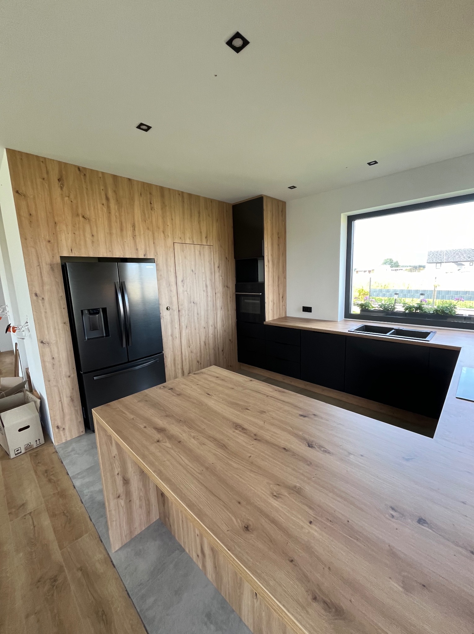 Nowoczesna kuchnia z drewnianymi frontami i czarnymi akcentami, duża wyspa kuchenna z blatem o naturalnym wykończeniu, czarna lodówka side-by-side wkomponowana w zabudowę, duże okno z widokiem...