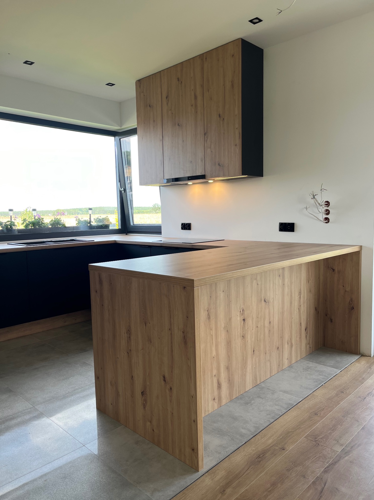 Nowoczesna kuchnia z drewnopodobnymi frontami szafek wiszących i półwyspem, czarne dolne szafki, duże okno z widokiem na zieleń, widoczne niedokończone instalacje elektryczne.