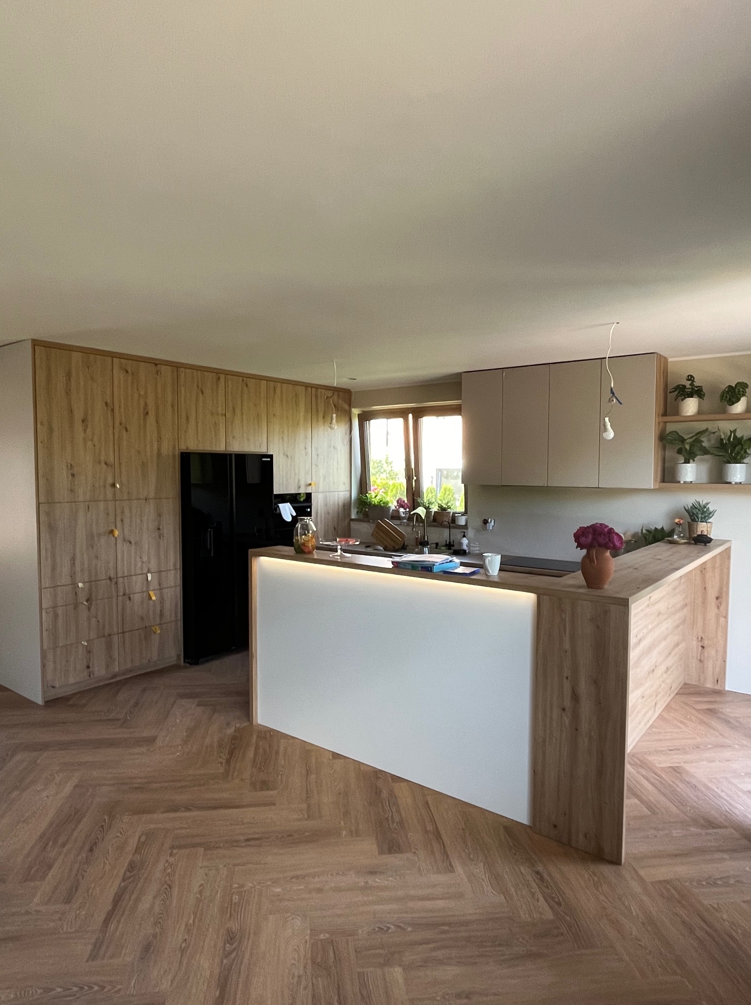 Nowoczesna kuchnia z drewnianymi szafkami, wyspą z podświetleniem LED i parkietem w jodełkę, czarna lodówka w zabudowie.