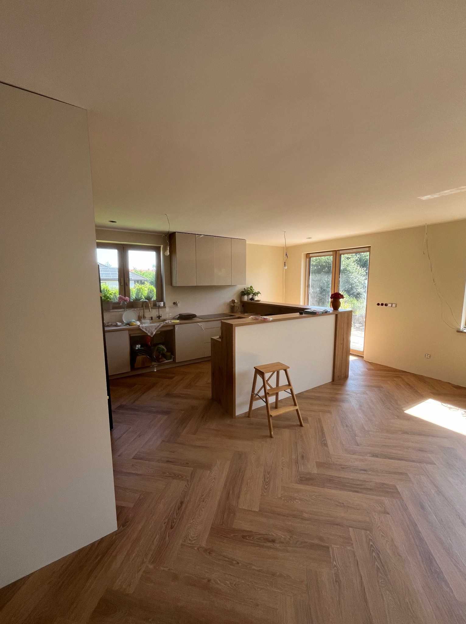 Wnętrze w trakcie wykańczania: jasna kuchnia z wyspą, drewniana podłoga ułożona w jodełkę, widoczne niedokończone instalacje elektryczne i drabina malarska.