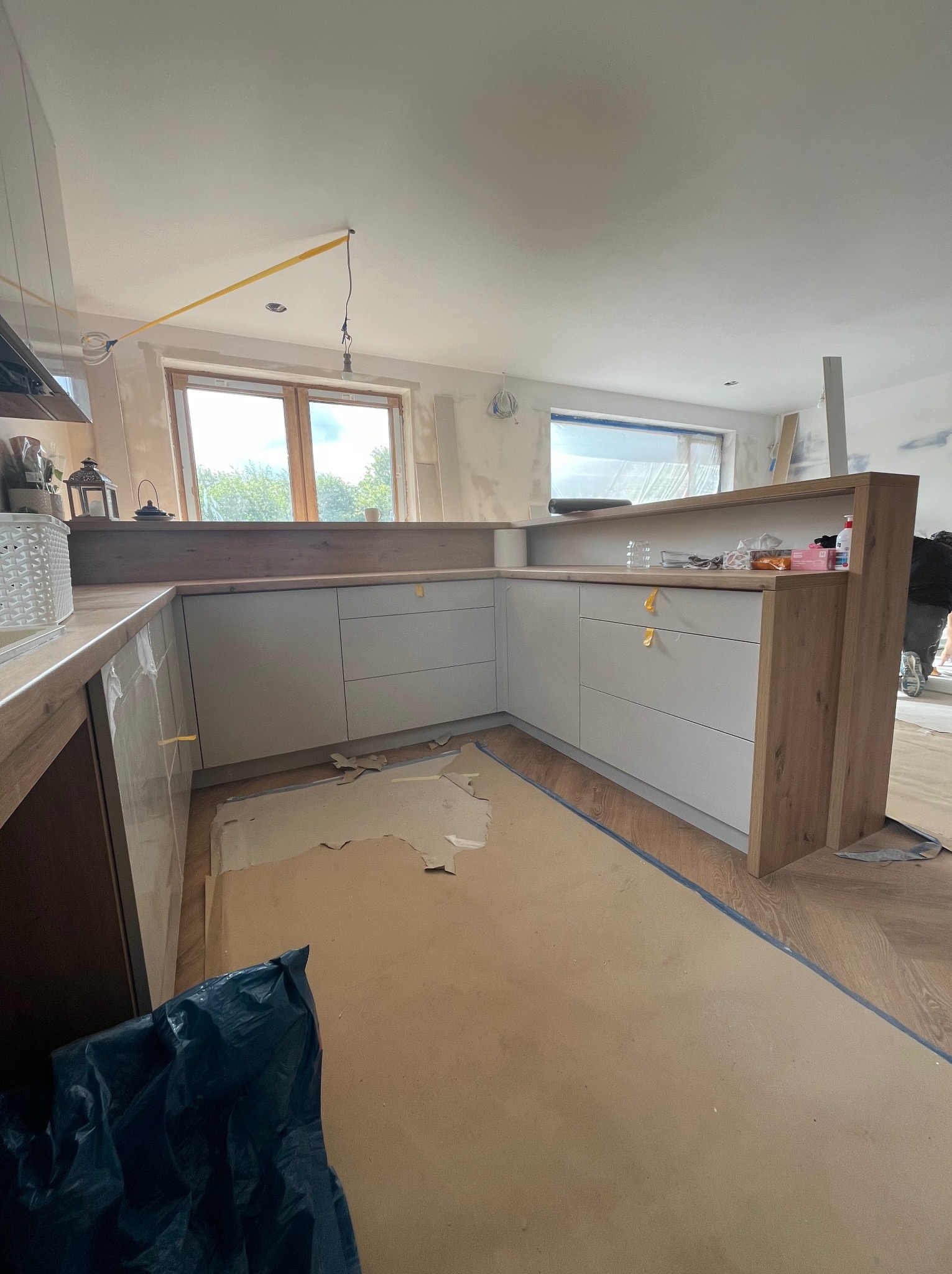 Montaż szarej, narożnej zabudowy kuchennej z drewnianymi elementami wykończeniowymi, widoczne zabezpieczenia podłogi kartonem i folią, surowy stan ścian, okno w tle.