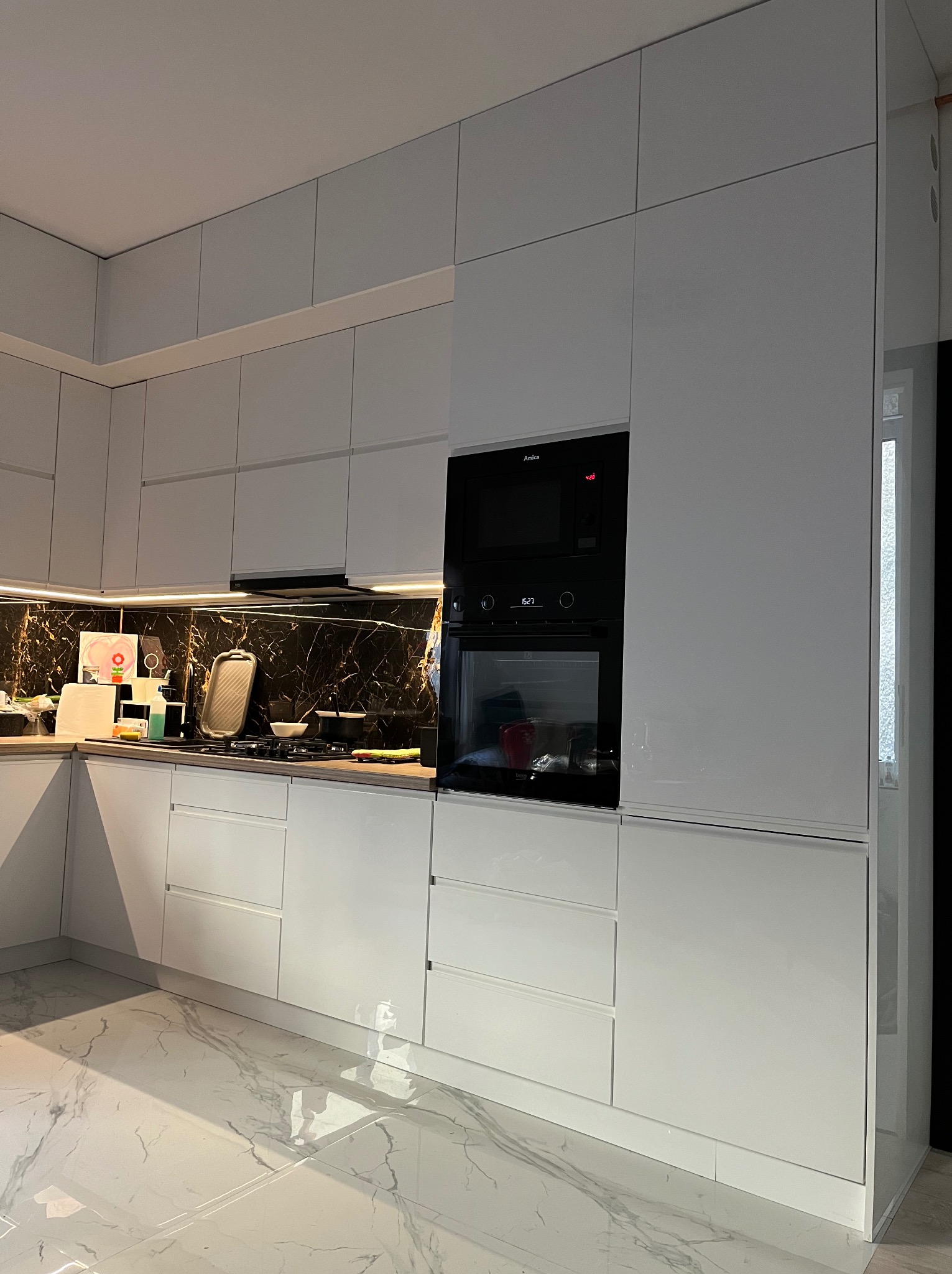 Biała, nowoczesna kuchnia z połyskiem, ciemny marmurowy backsplash podświetlony listwą LED, sprzęt AGD marki Amica, podłoga z białego marmuru z szarymi żyłami.