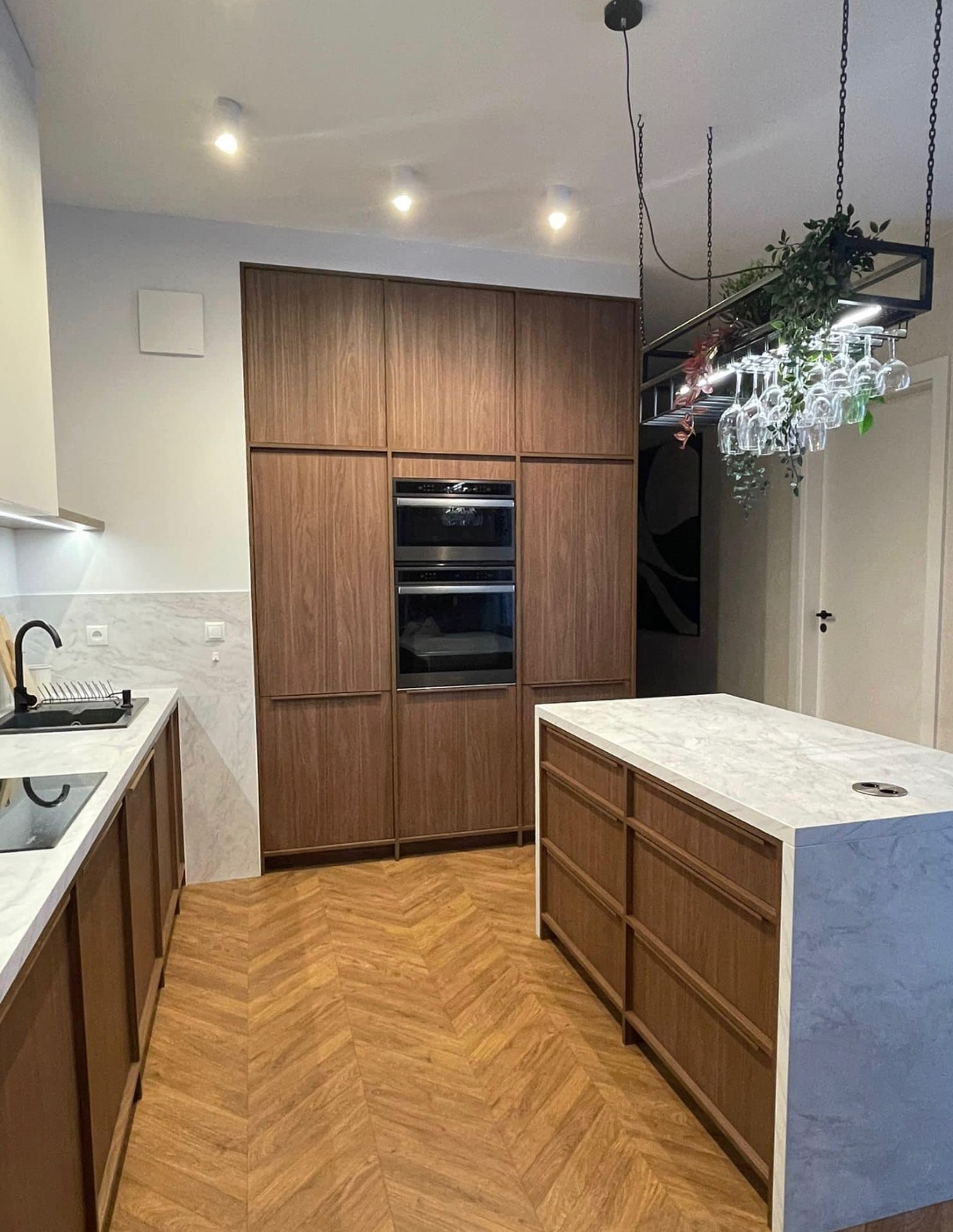 Nowoczesna kuchnia z drewnianymi frontami szafek, marmurowym blatem, wyspą kuchenną i podłogą ułożoną w jodełkę. Nad wyspą wisi czarna konstrukcja z roślinami i kieliszkami.