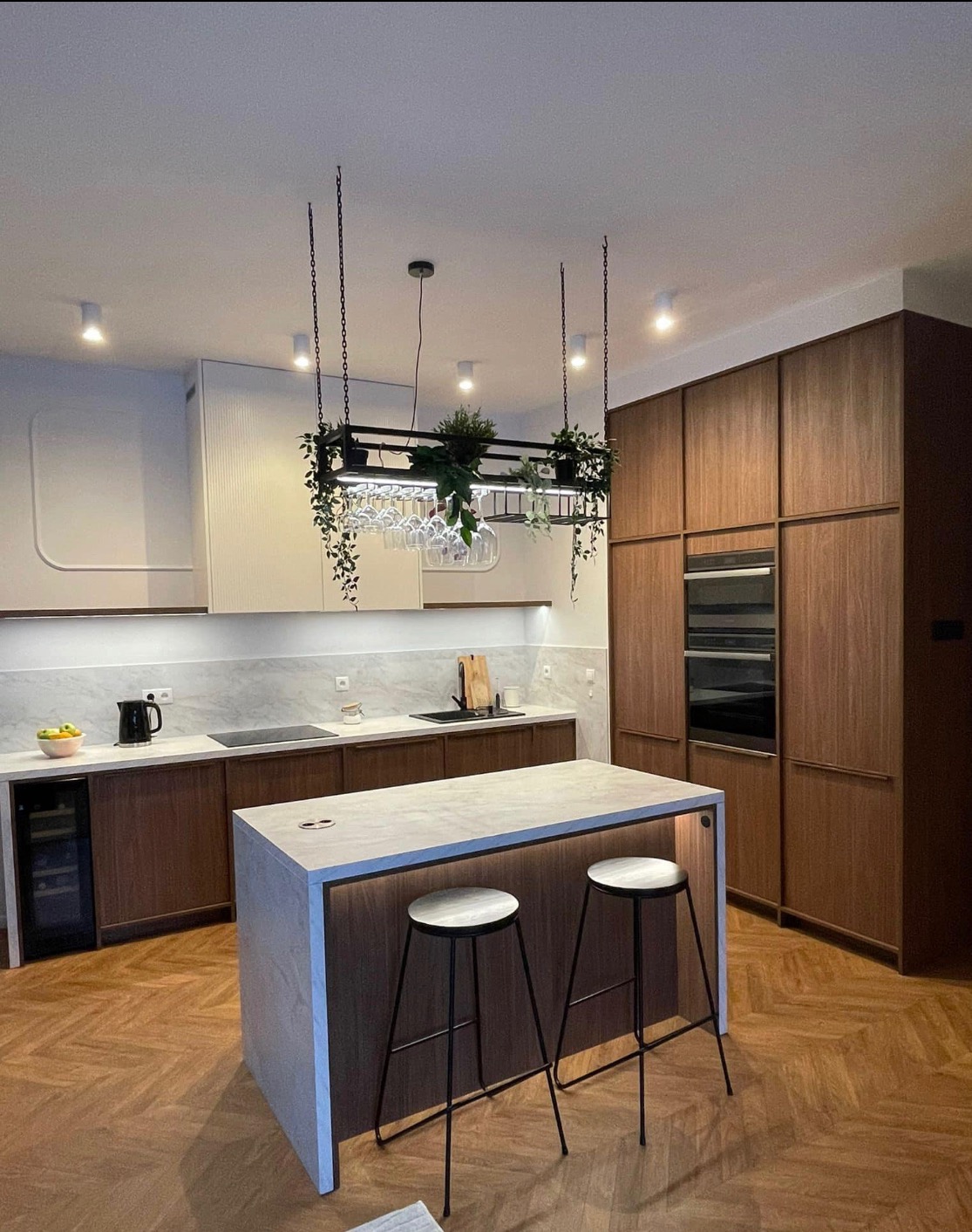 Nowoczesna kuchnia z wyspą oświetloną od dołu, drewnianymi frontami szafek, marmurowym blatem, wiszącym regałem na kieliszki z roślinami i parkietem ułożonym w jodełkę.