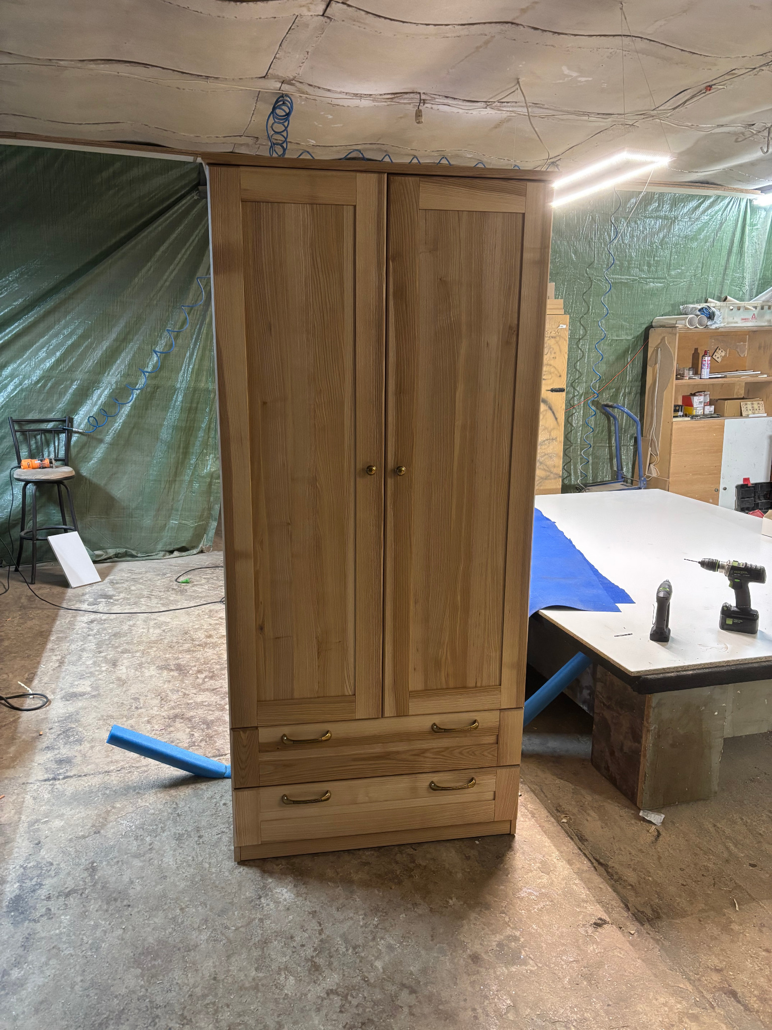 Dwudrzwiowa, drewniana szafa z dwiema szufladami na tle warsztatu z narzędziami i folią ochronną.