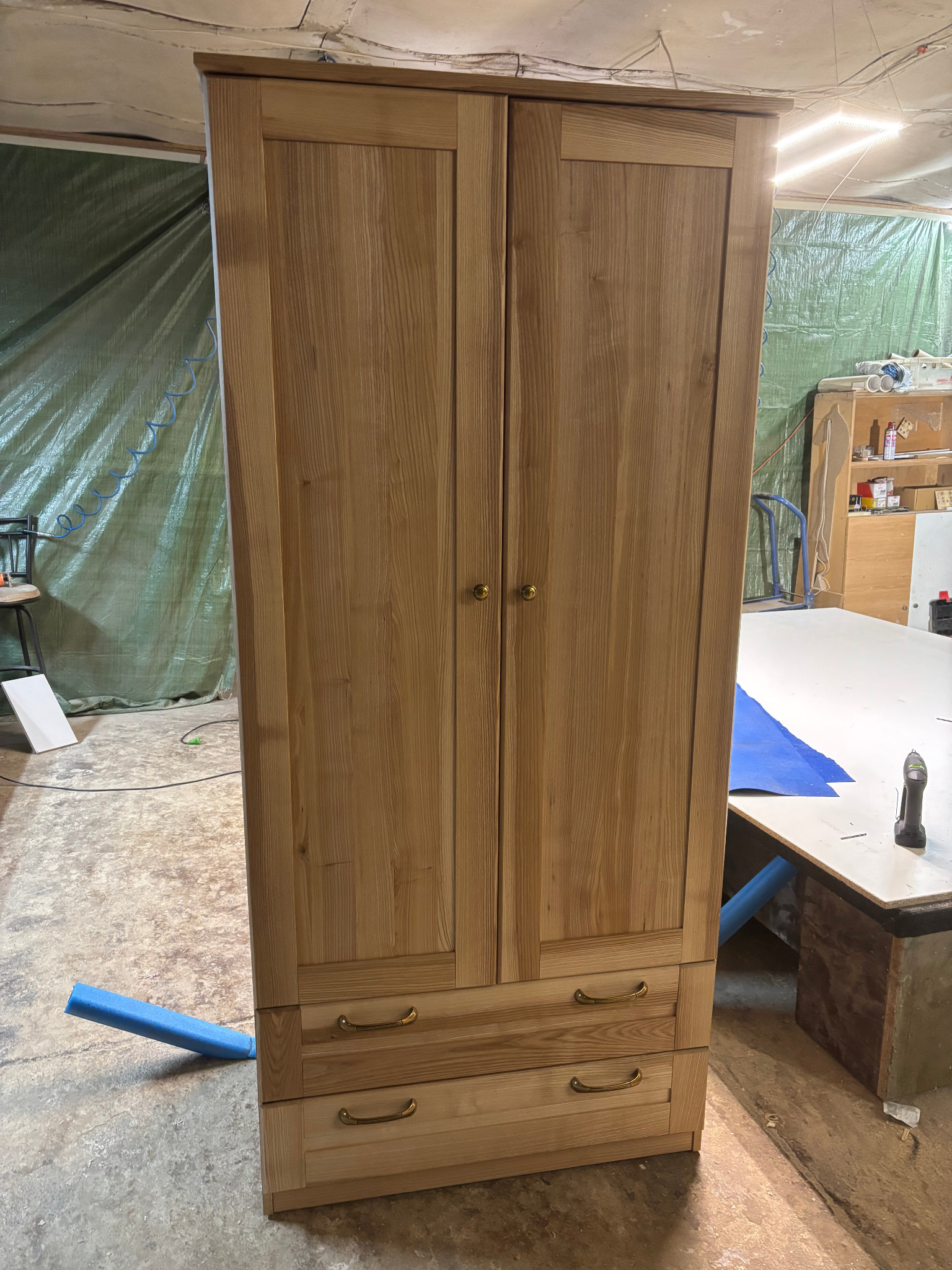 Dębowa szafa dwudrzwiowa z dwiema szufladami, wykończona złotymi gałkami i uchwytami, sfotografowana w warsztacie na tle zielonej folii ochronnej.