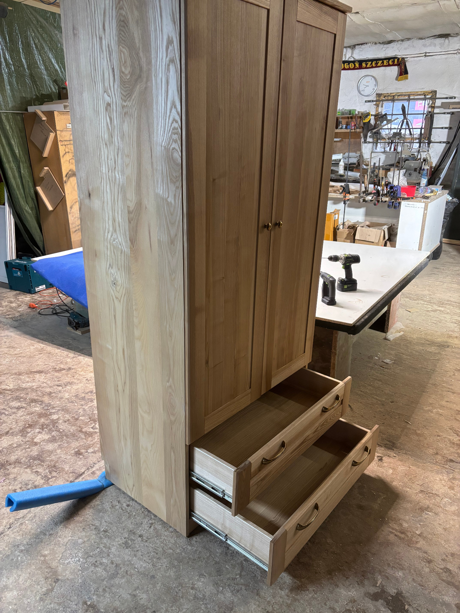 Wysoka, dwudrzwiowa szafa z jasnego drewna z dwoma wysuwanymi szufladami, widoczna w warsztacie, z wiertarką i wkrętarką akumulatorową leżącą na stole obok.