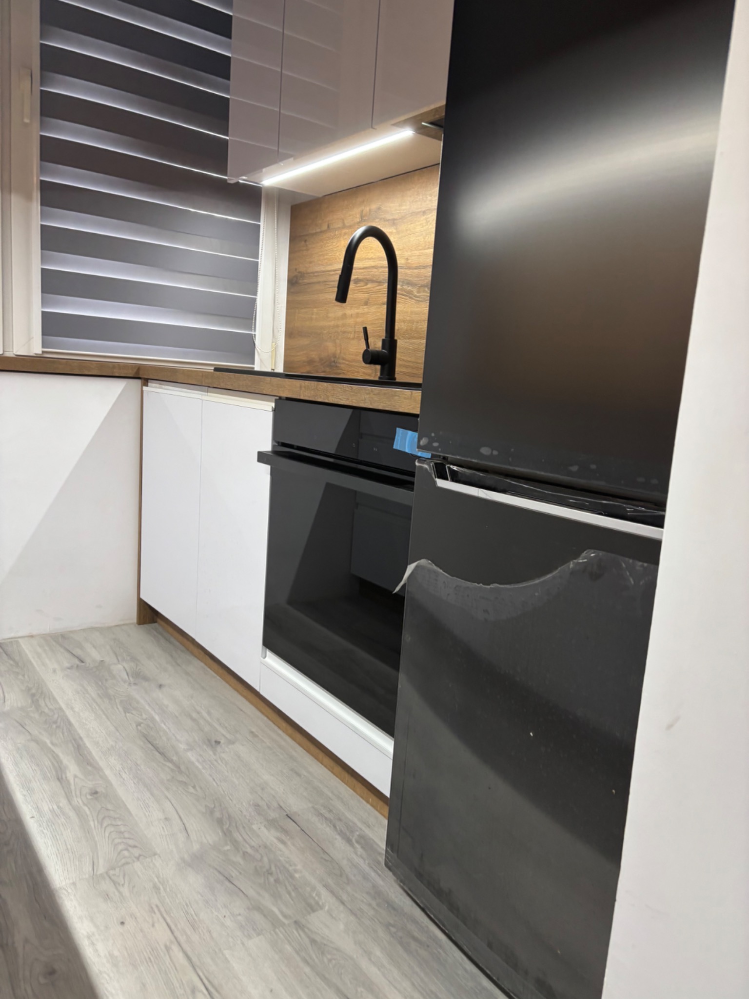 Nowoczesna kuchnia: białe szafki z drewnianym blatem, czarna armatura i piekarnik. Widoczny fragment lodówki z folią ochronną. Żaluzje okienne w tle.