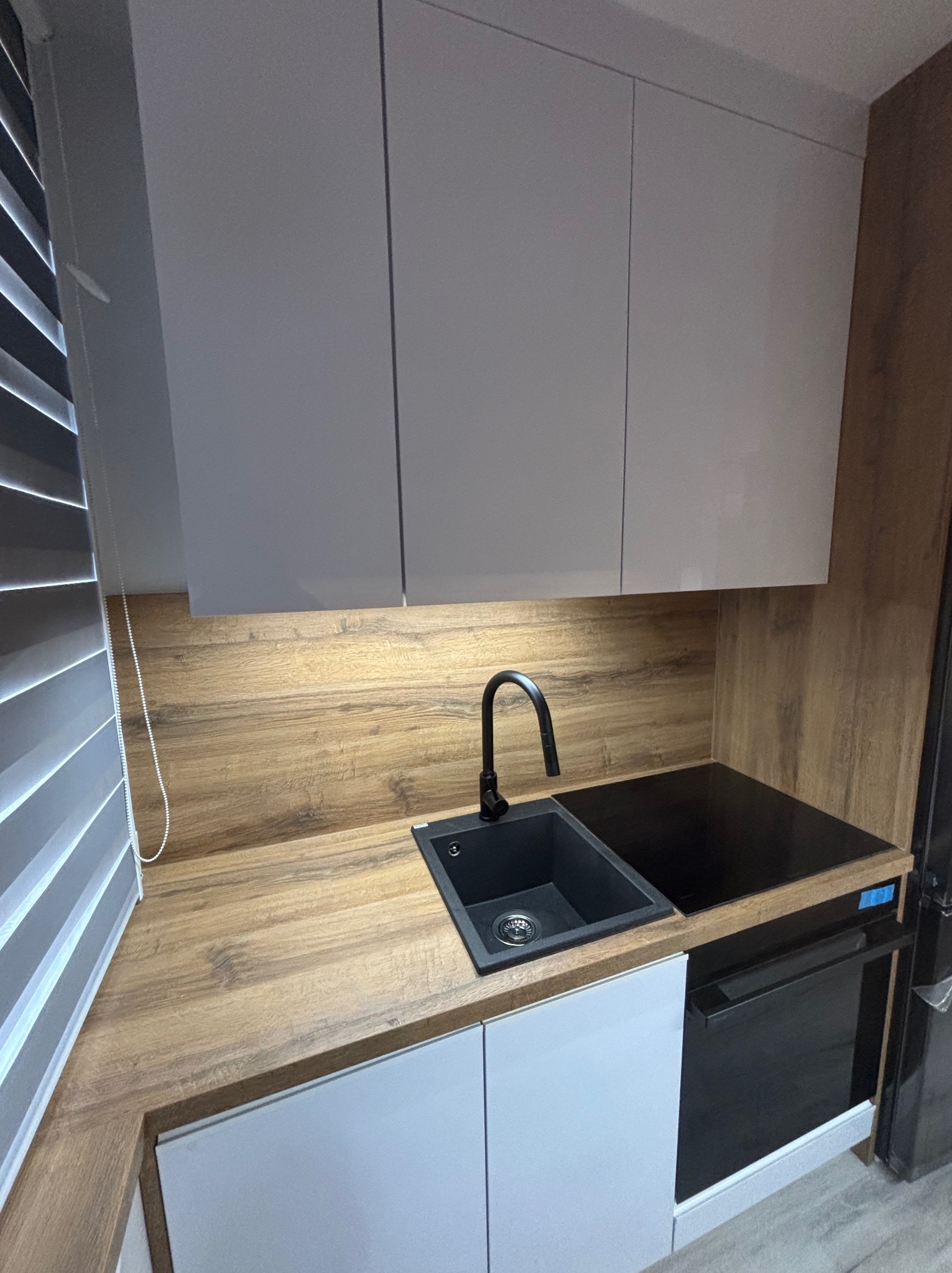 Nowoczesna kuchnia: szare szafki, drewniany blat, czarny zlew i bateria, płyta indukcyjna. Minimalistyczny design z żaluzjami w oknie.