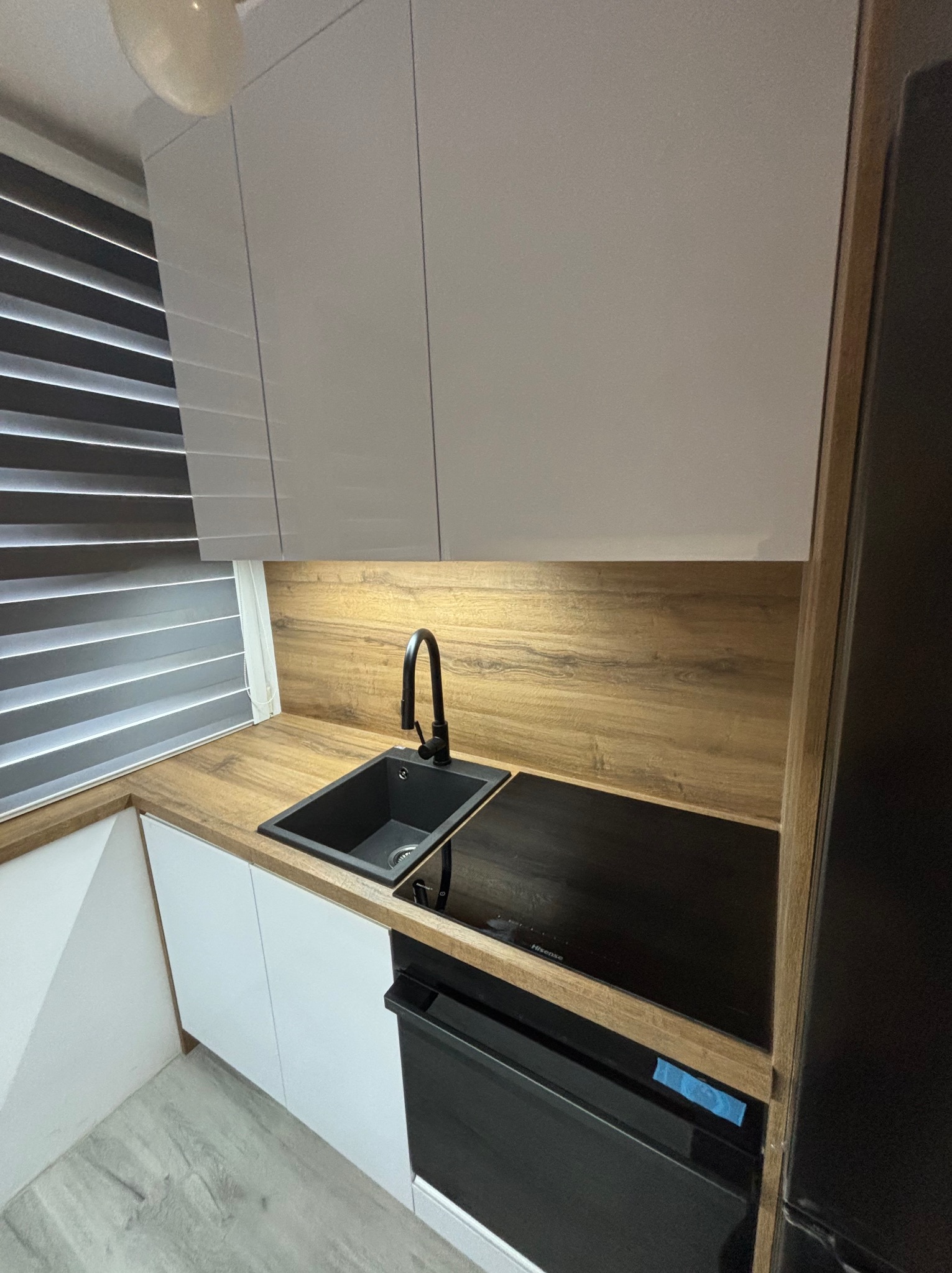 Nowoczesna kuchnia: białe szafki wiszące, drewniany blat, czarny zlew i płyta indukcyjna. Minimalistyczny design z żaluzjami w oknie.