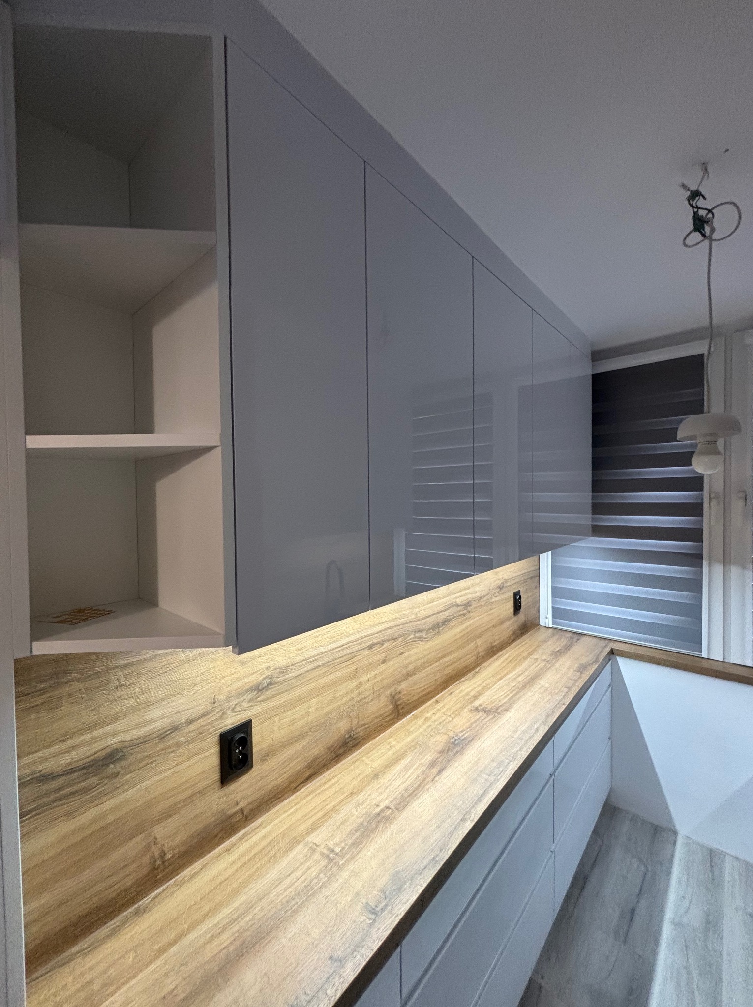 Nowoczesna kuchnia z szarymi szafkami i drewnianym blatem. Widoczne gniazdko elektryczne w blacie i otwarte półki. Minimalistyczny design i roleta w oknie.