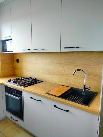 Jasna kuchnia na wymiar: białe szafki z czarnymi uchwytami, drewniany blat, zlew z baterią i płyta gazowa. Minimalistyczny design z drewnianym akcentem.