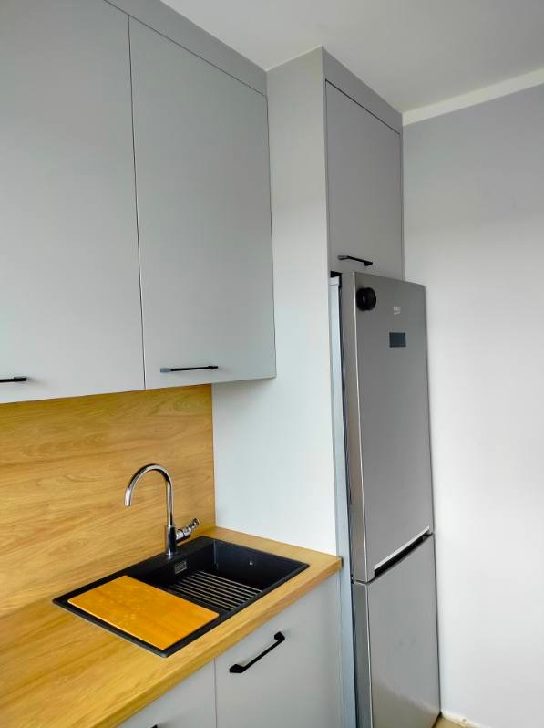 Szare szafki kuchenne z drewnianym blatem, czarnym zlewozmywakiem i srebrną lodówką. Minimalistyczny design, nowoczesne wykończenie.