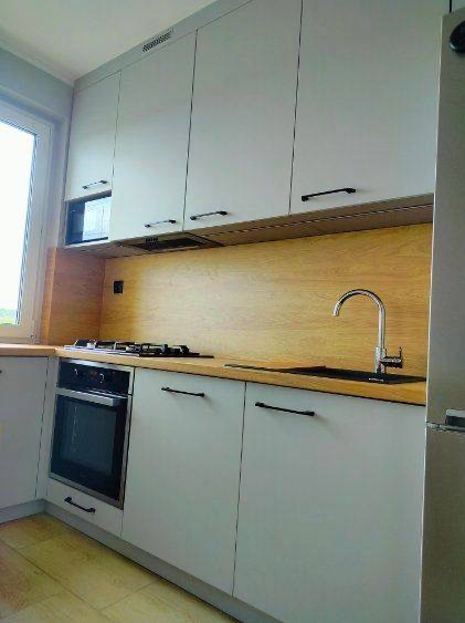Jasna kuchnia z drewnianym blatem i czarnymi uchwytami. Widoczny zlew, płyta grzewcza i piekarnik. Minimalistyczny design z gładkimi frontami szafek.