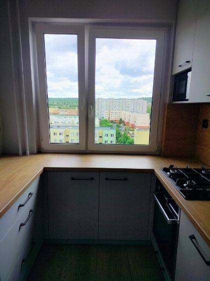 Nowoczesna kuchnia z oknem i widokiem na miasto. Jasne fronty szafek, drewniany blat i płyta grzewcza. Minimalistyczny design z akcentem na funkcjonalność.