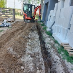 D.W prace ziemne i ogrodowe - Wąski wykop pod fundament domu z bloczków, wykonany pomarańczową minikoparką Kubota. Widoczne świeże ślady opon i ułożone bloczki na paletach. Tło: brzozy i drewniane ogrodzenie.