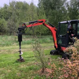 Czerwona minikoparka Kubota z wiertłem w trawiastym terenie, operator w kabinie. W tle drzewa, z przodu krzew z różowymi liśćmi. Wykonanie wykopu pod nasadzenia.
