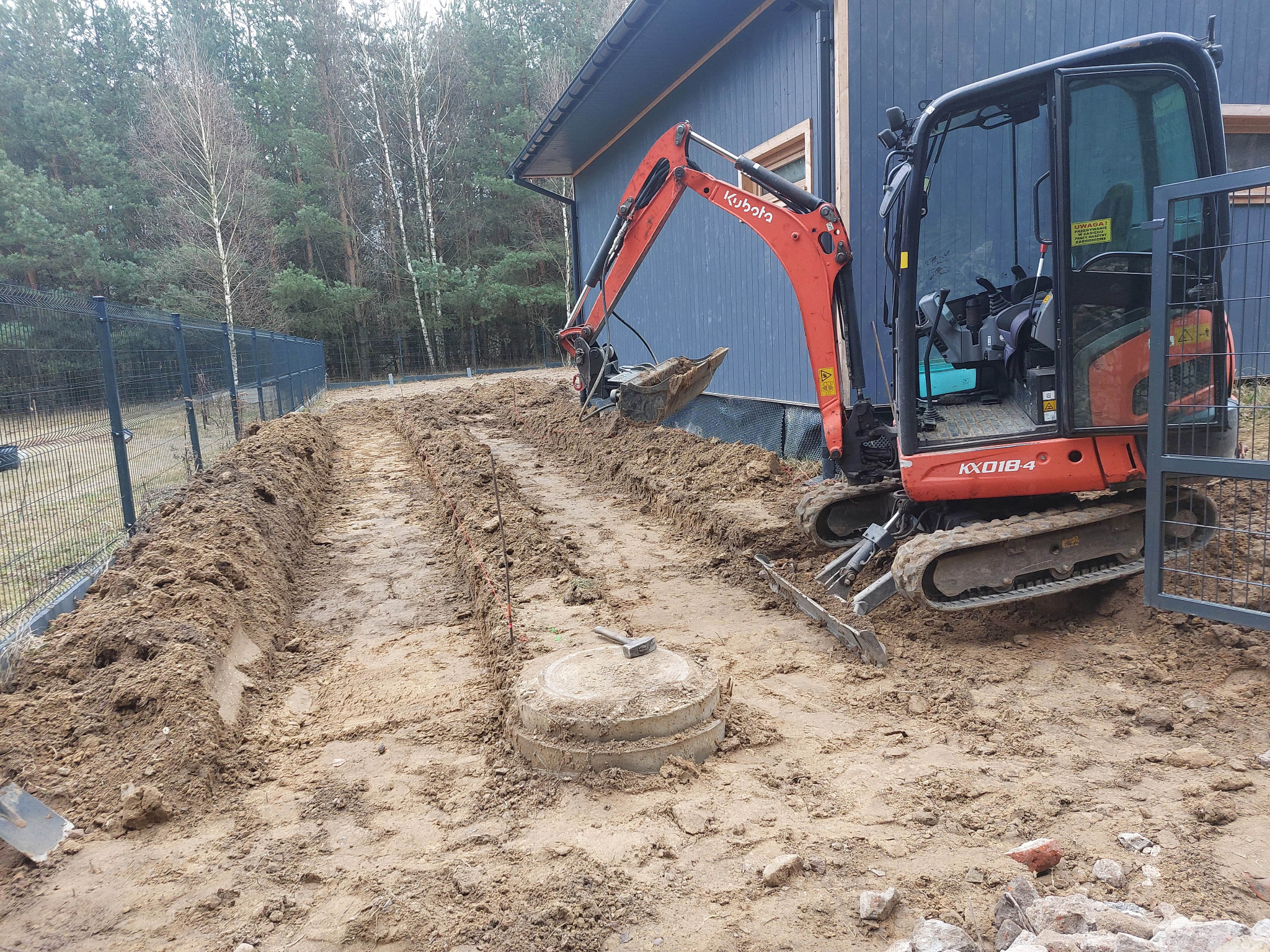 Pomarańczowa minikoparka Kubota przy wykopie w ziemi, obok betonowa studzienka. W tle budynek z ciemnego drewna i ogrodzenie siatkowe.
