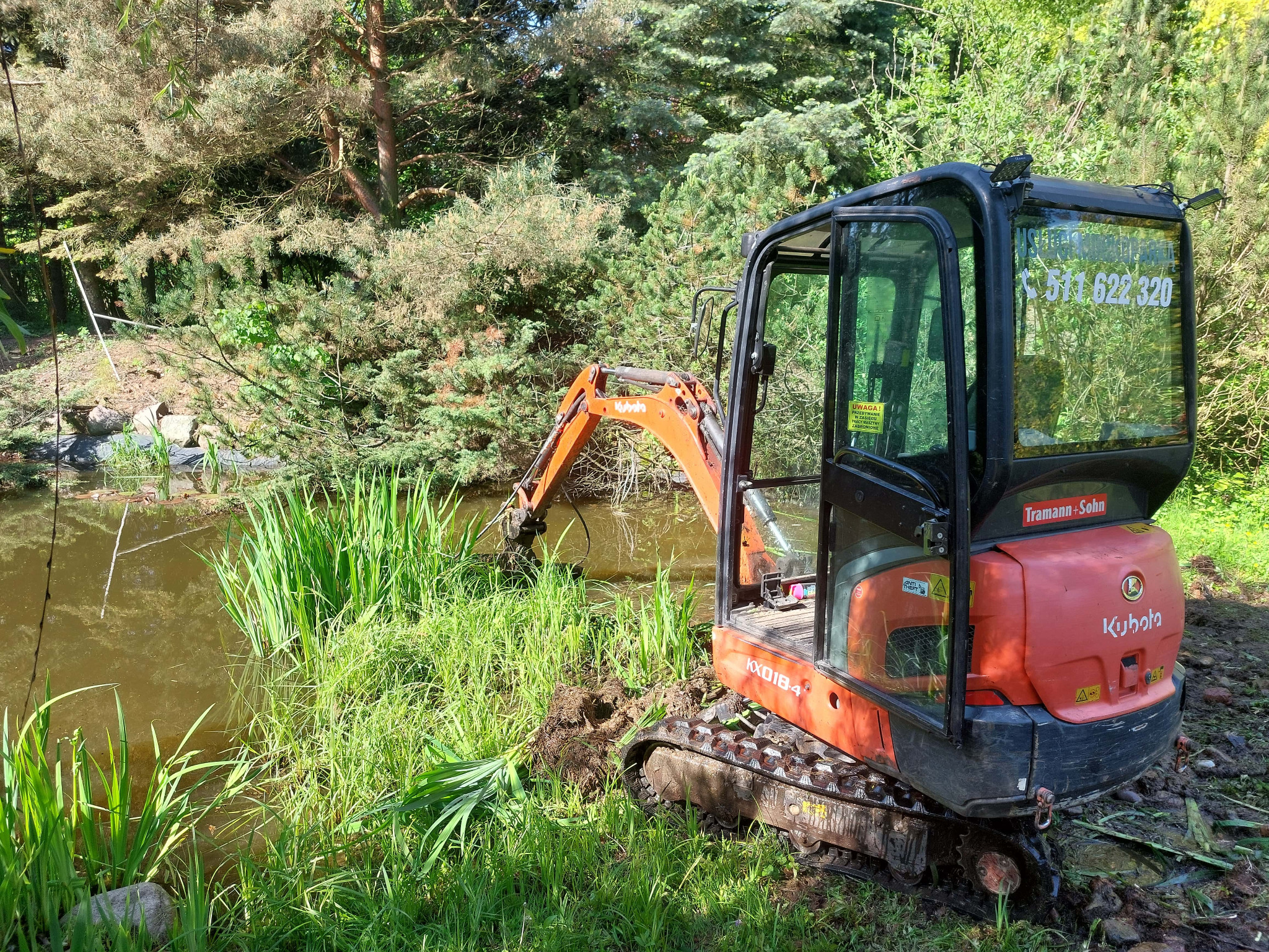 Pomarańczowa minikoparka Kubota KX018-4, z otwartą kabiną, częściowo zanurzona w stawie z roślinnością, na tle drzew i krzewów, z numerem telefonu na szybie.