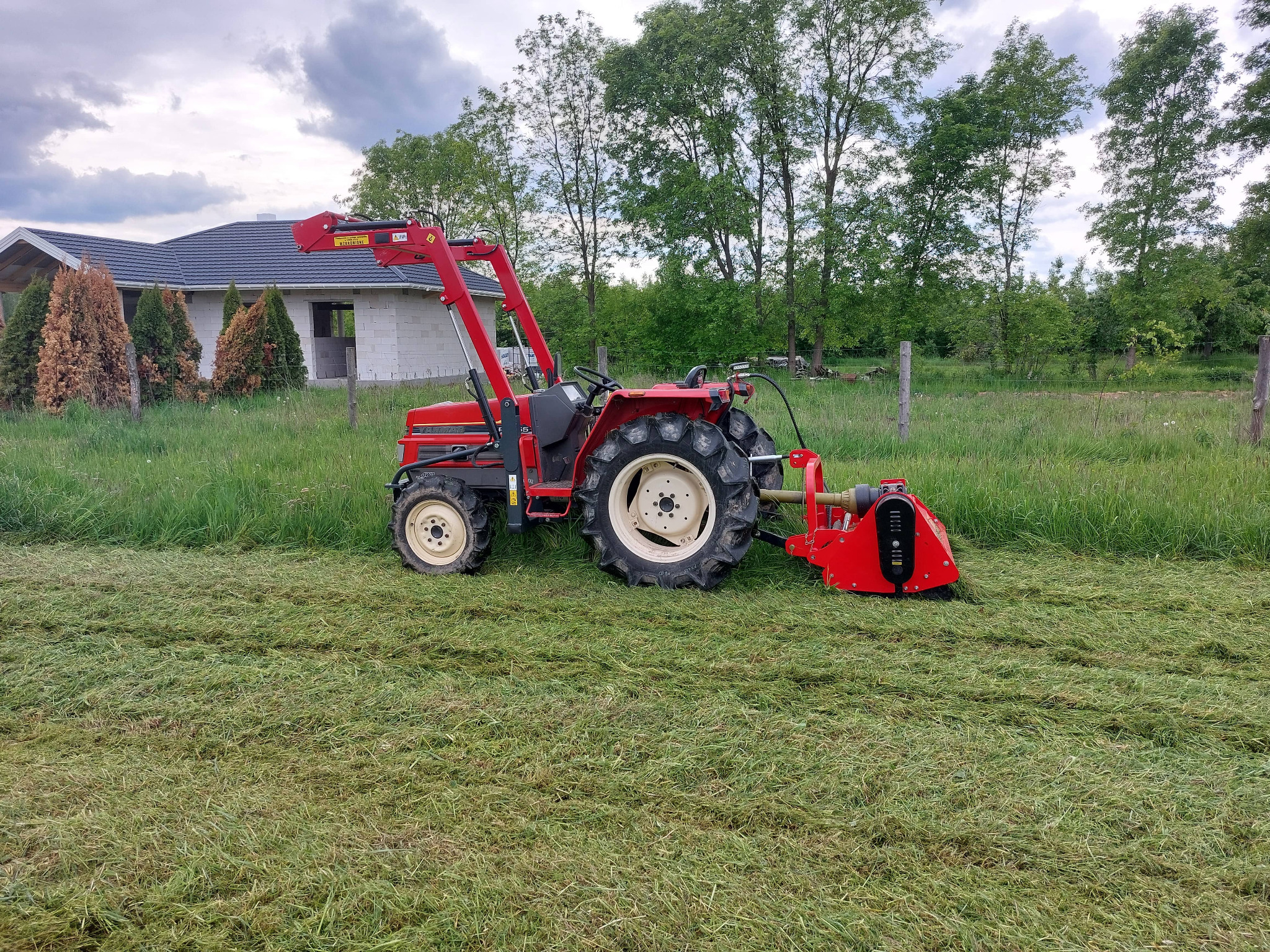 Czerwony traktor z kosiarką rotacyjną na polu po koszeniu trawy. W tle dom w budowie i drzewa. Widoczne ślady koszenia.
