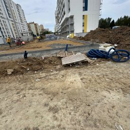 ELEKTROBUD Sp. z o.o. - Teren budowy z widocznymi rurami karbowanymi, paletą, stertą ziemi i fragmentami fundamentów na tle nowych bloków mieszkalnych.