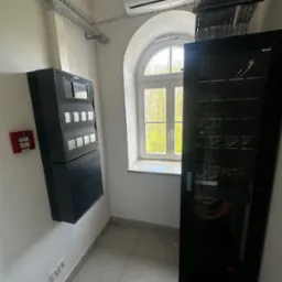 Centrala alarmowa Bosch AVENAR i szafa rack z okablowaniem sieci LAN w pomieszczeniu z oknem łukowym i szynami kablowymi pod sufitem.