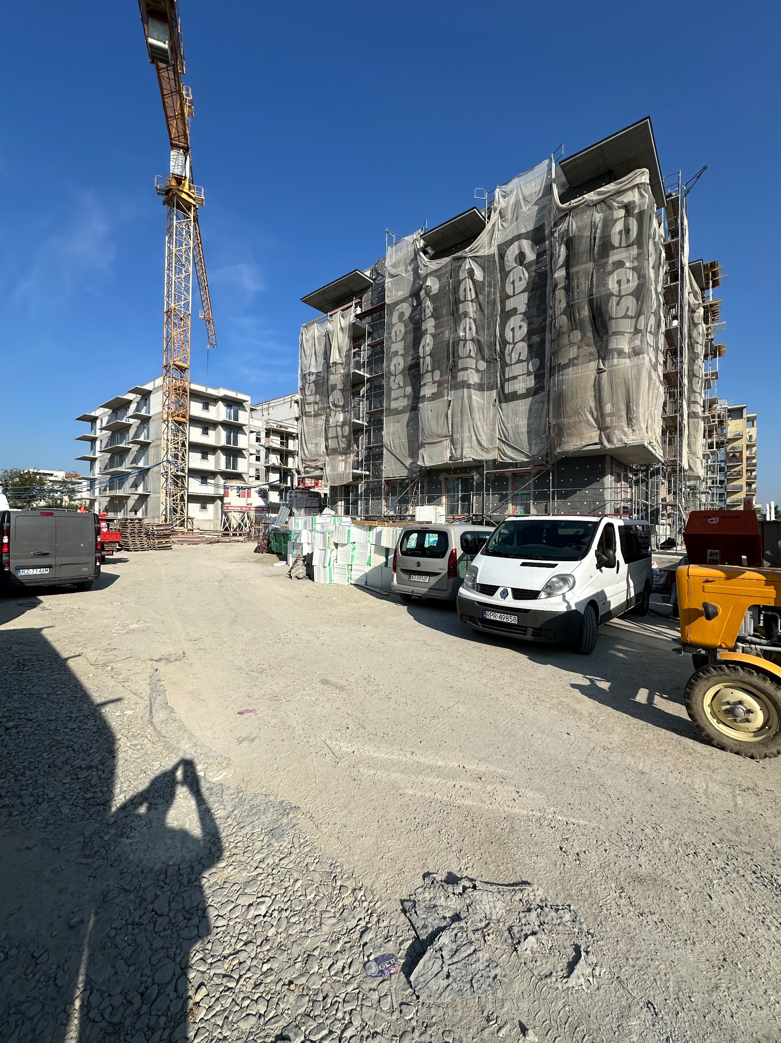 Budynek w trakcie ocieplania, osłonięty siatką z logo producenta, widoczny dźwig budowlany, zaparkowane busy i traktor.