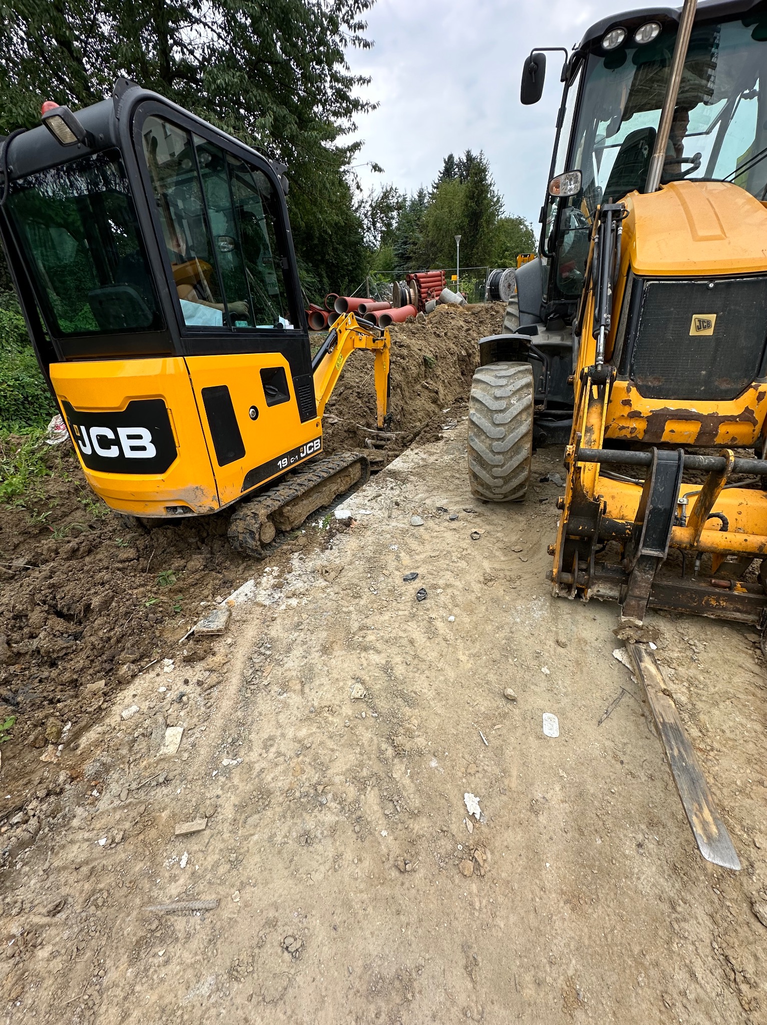 Żółty minikoparka JCB 19C-1 i traktor JCB z łyżką kopią w ziemi, w tle rury i zieleń.