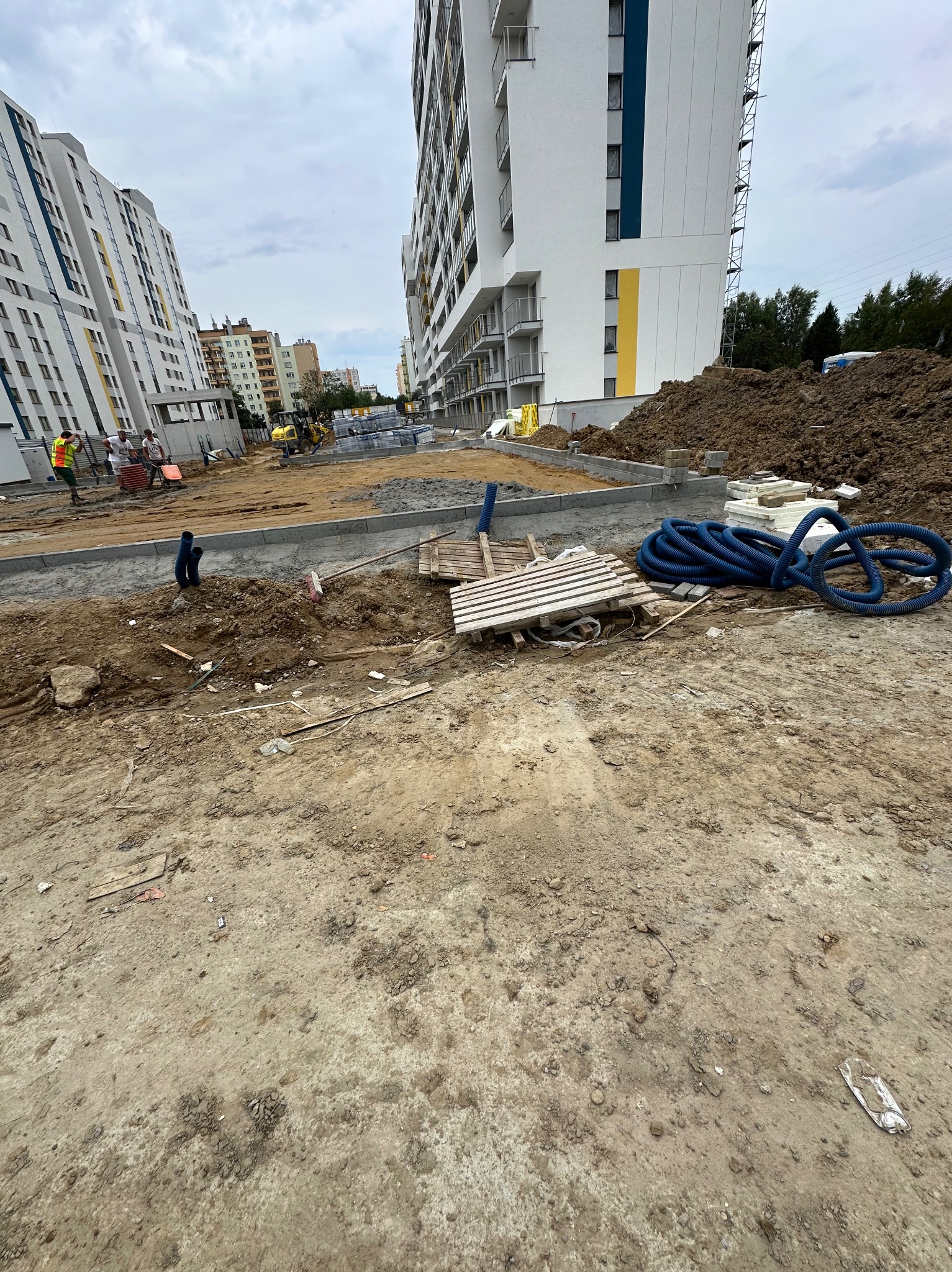 Teren budowy z widocznymi rurami karbowanymi, paletą, stertą ziemi i fragmentami fundamentów na tle nowych bloków mieszkalnych.