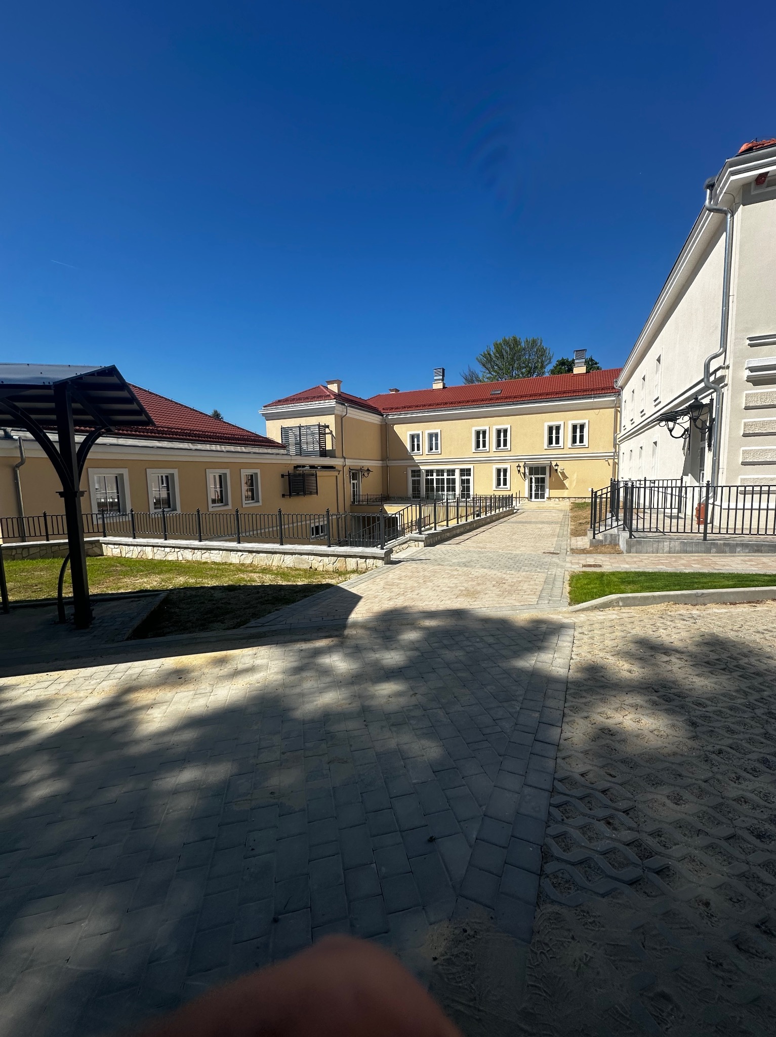 Elewacja budynku w odcieniach beżu z czerwonym dachem, czarne metalowe balustrady i oświetlenie zewnętrzne, kostka brukowa na podjeździe, błękitne niebo.