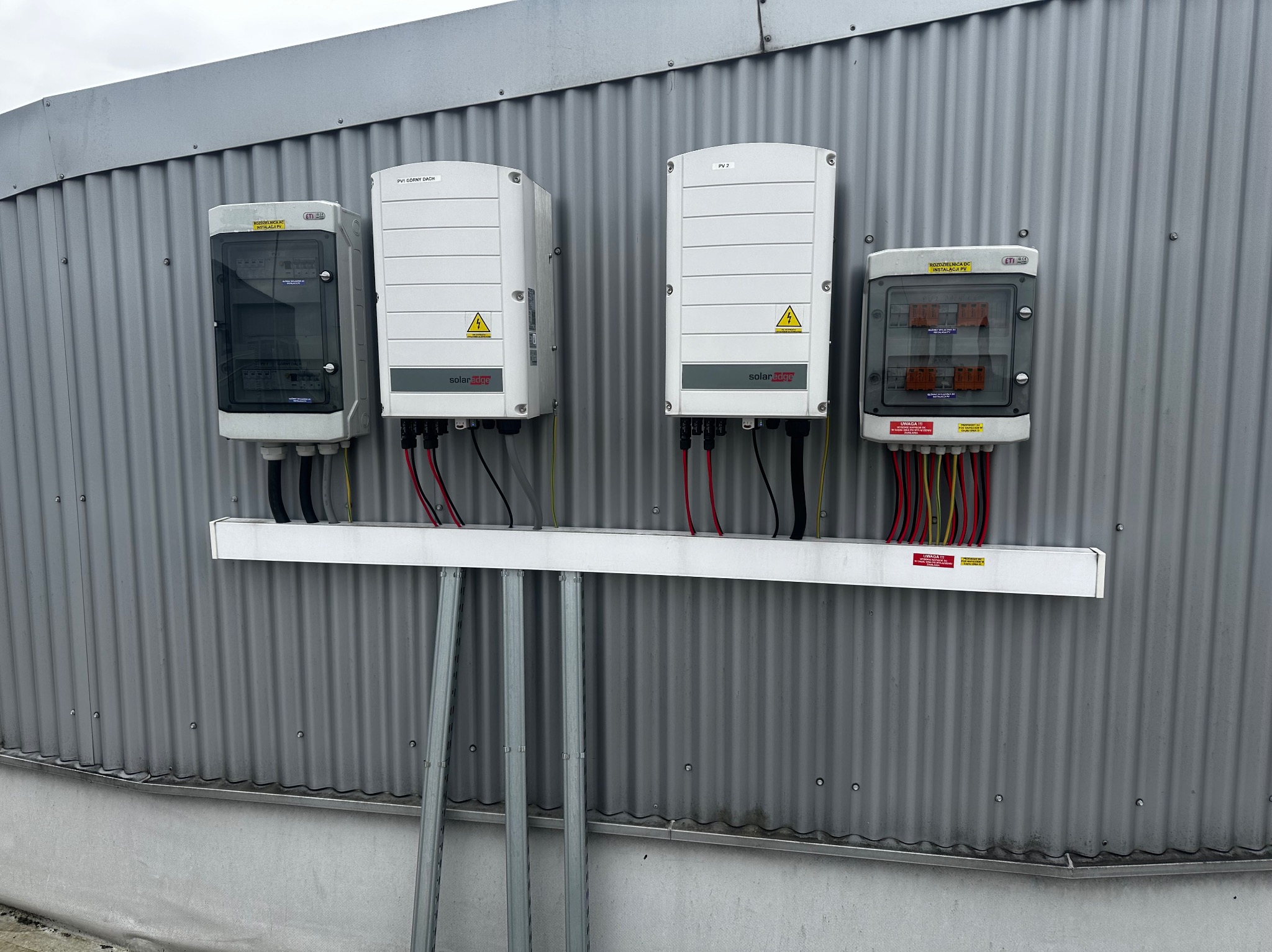 Rozdzielnice elektryczne DC i inwertery solarne zamontowane na falistej ścianie z blachy trapezowej, kable prowadzone w rurach osłonowych.