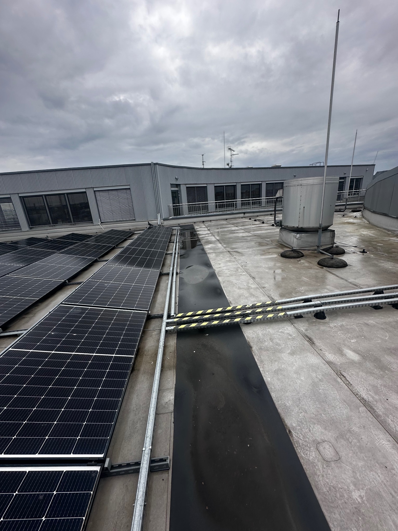 Widok z góry na panele słoneczne zamontowane na dachu przemysłowego budynku w pochmurny dzień. Widoczne metalowe konstrukcje wsporcze, elementy wentylacji i instalacje odgromowe.