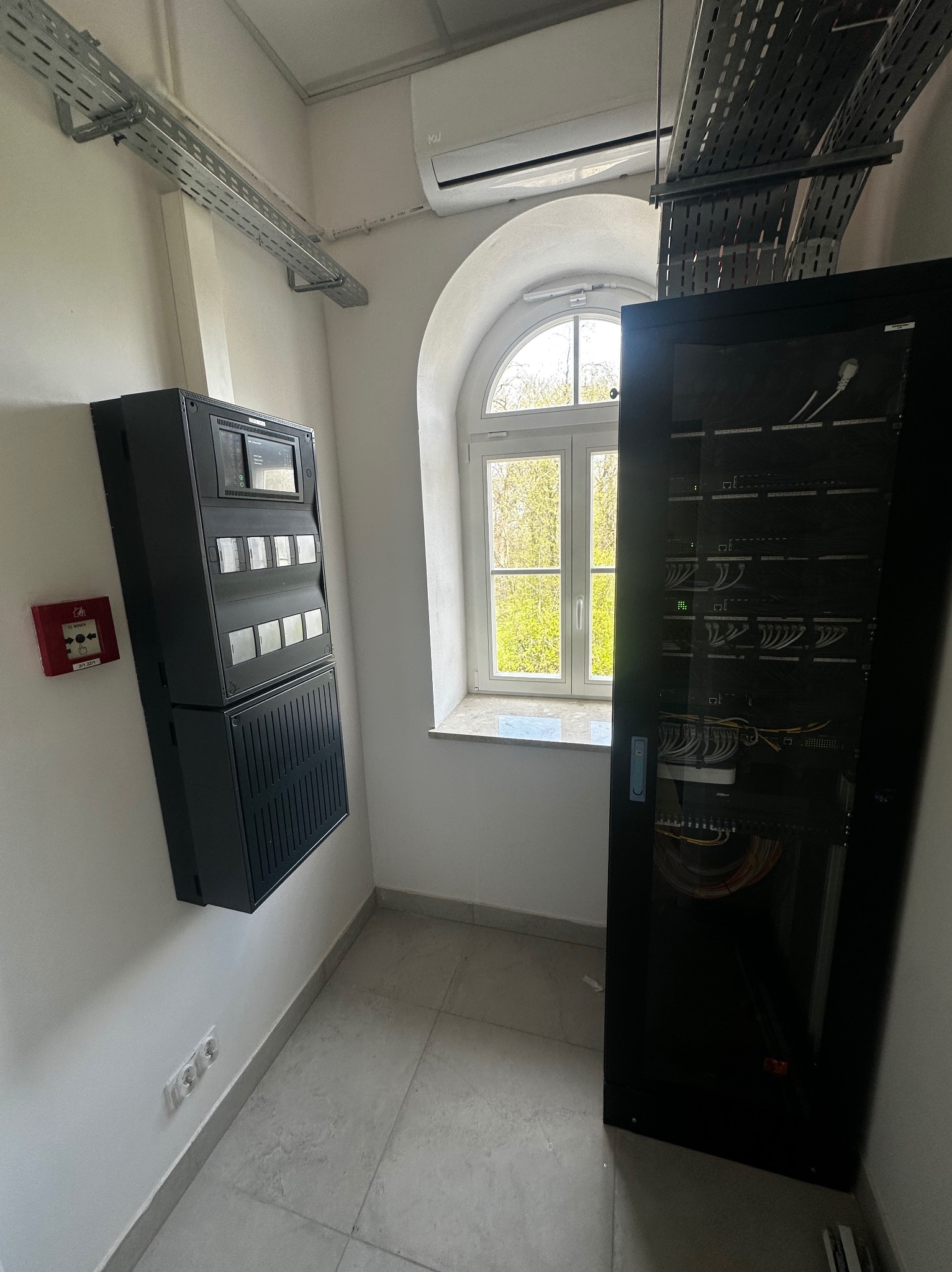 Centrala alarmowa Bosch AVENAR i szafa rack z okablowaniem sieci LAN w pomieszczeniu z oknem łukowym i szynami kablowymi pod sufitem.