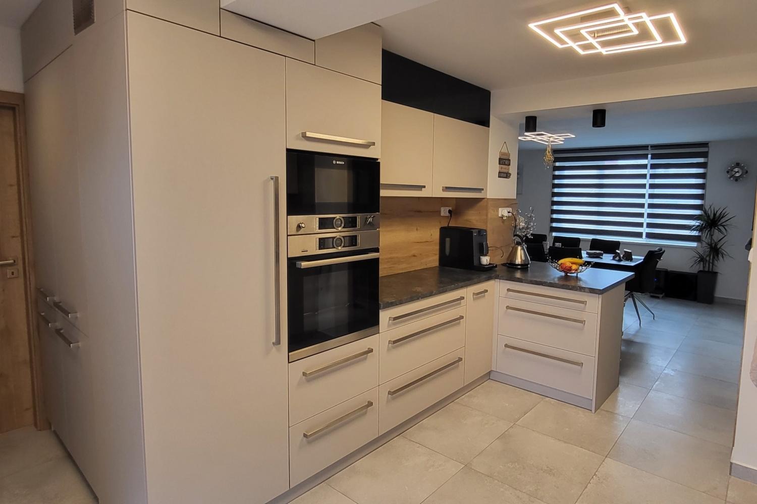 Jasna kuchnia z zabudową, piekarnikiem i mikrofalówką w słupku, szufladami z długimi uchwytami, drewnianym panelem nad blatem i nowoczesnym oświetleniem sufitowym.