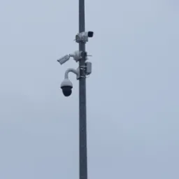 Słup z trzema kamerami monitoringu różnego typu, skierowanymi w różnych kierunkach, na tle pochmurnego nieba.