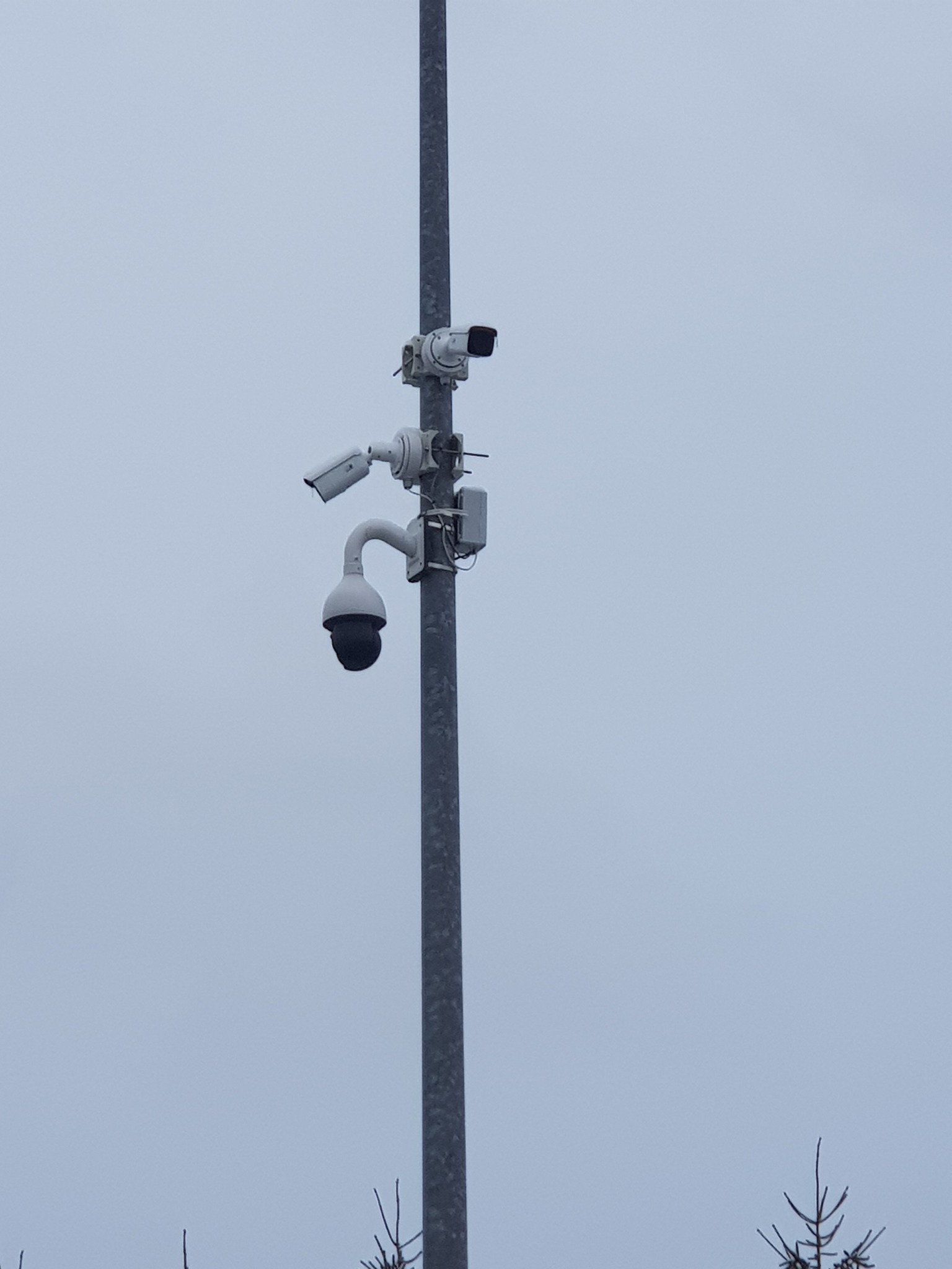 Słup z trzema kamerami monitoringu różnego typu, skierowanymi w różnych kierunkach, na tle pochmurnego nieba.