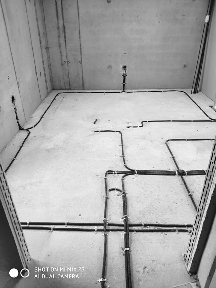 Surowe wnętrze w trakcie prac, widoczne kable elektryczne przymocowane do surowego betonu ścian i podłogi za pomocą metalowych uchwytów, z perspektywy wejścia do pomieszczenia.