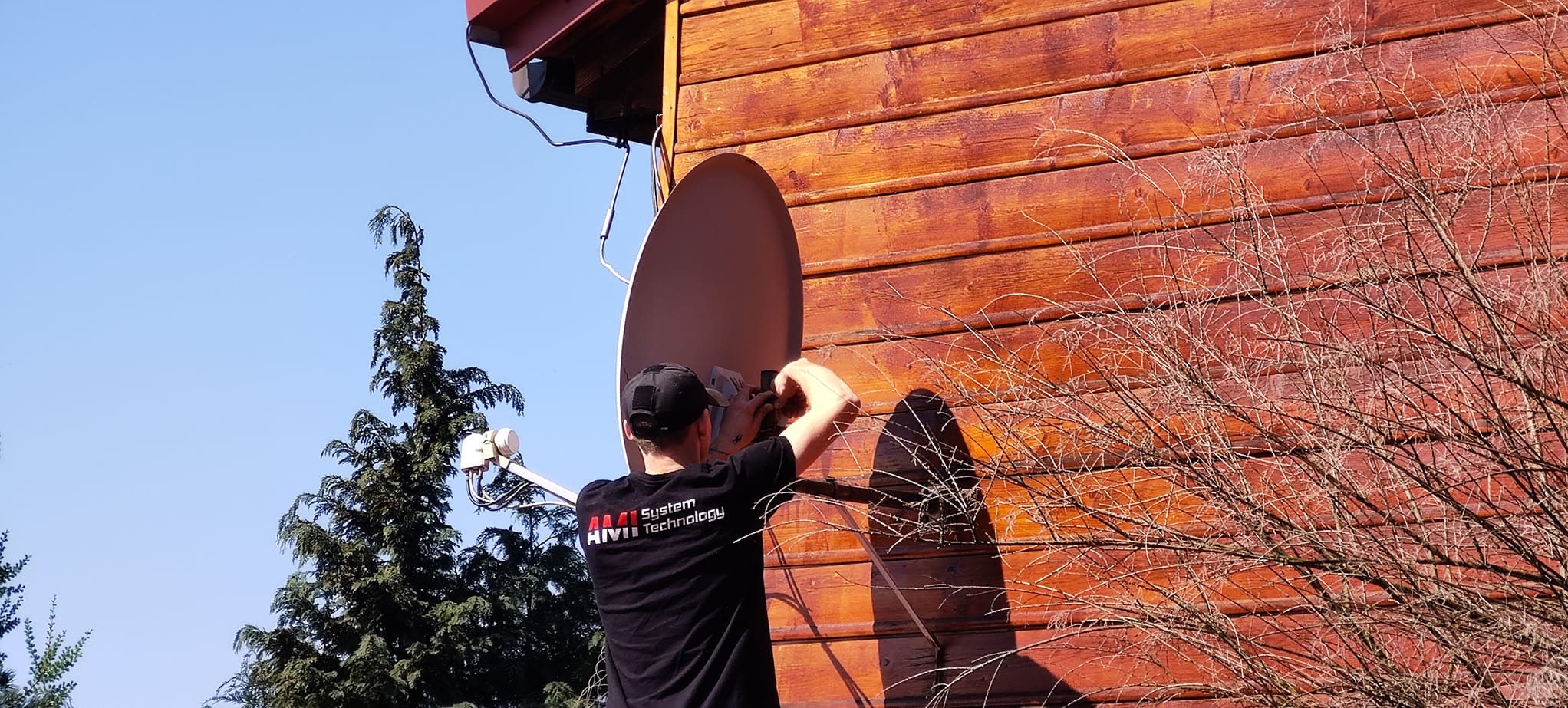 Instalator w czarnej koszulce z logo firmy montuje antenę satelitarną na drewnianej elewacji budynku, widoczny fragment drzewa iglastego i błękitne niebo.