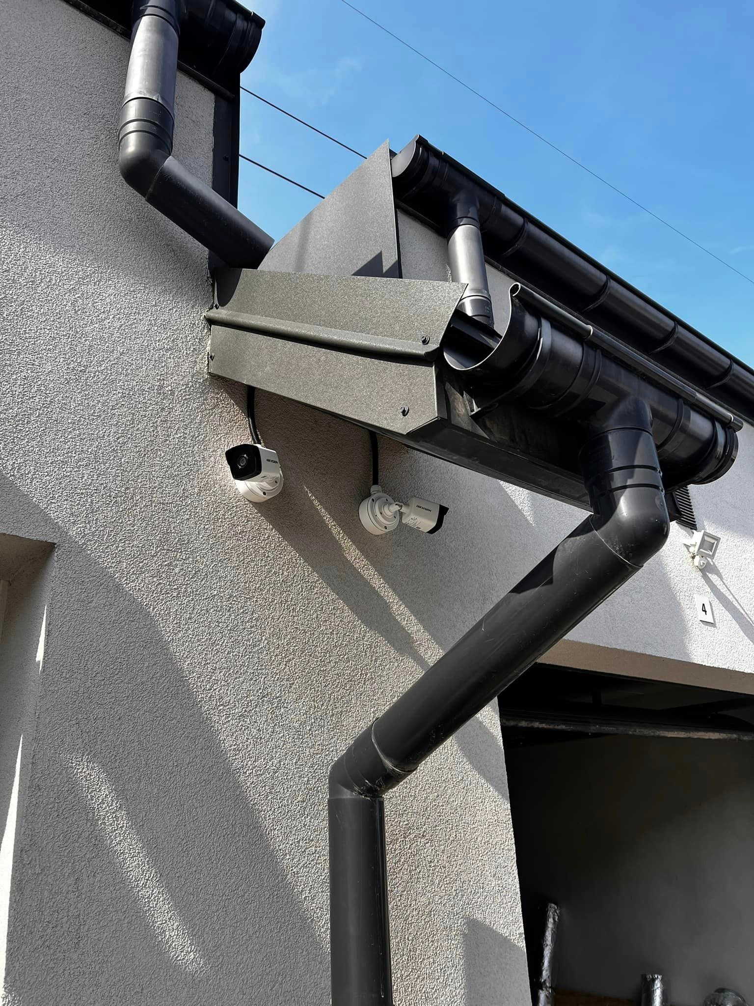 Dwie białe kamery monitoringu zamontowane pod okapem z czarnymi rynnami na elewacji budynku o szarej strukturze.