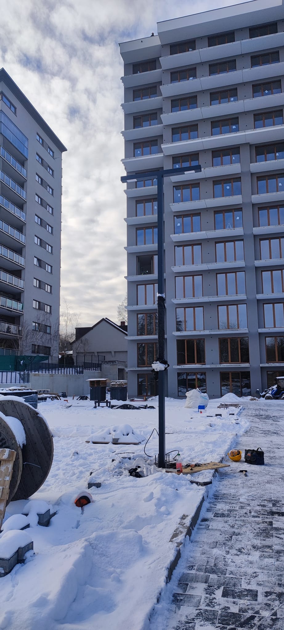 Montaż słupa oświetleniowego przed nowym budynkiem mieszkalnym w zimowej scenerii, widoczne narzędzia i kable elektryczne w śniegu.