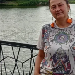 Kobieta z uśmiechem na twarzy, stojąca przy ozdobnym metalowym ogrodzeniu, z widokiem na rzekę i drzewa w tle, ubrana w koszulkę z kolorowym wzorem i szorty z ozdobnym paskiem.