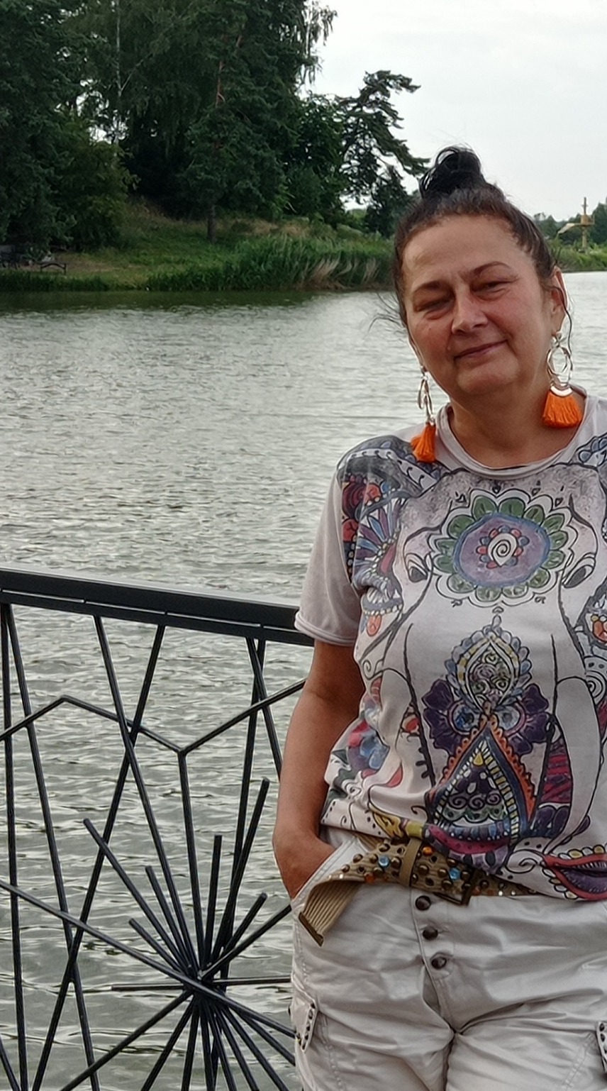 Kobieta z uśmiechem na twarzy, stojąca przy ozdobnym metalowym ogrodzeniu, z widokiem na rzekę i drzewa w tle, ubrana w koszulkę z kolorowym wzorem i szorty z ozdobnym paskiem.