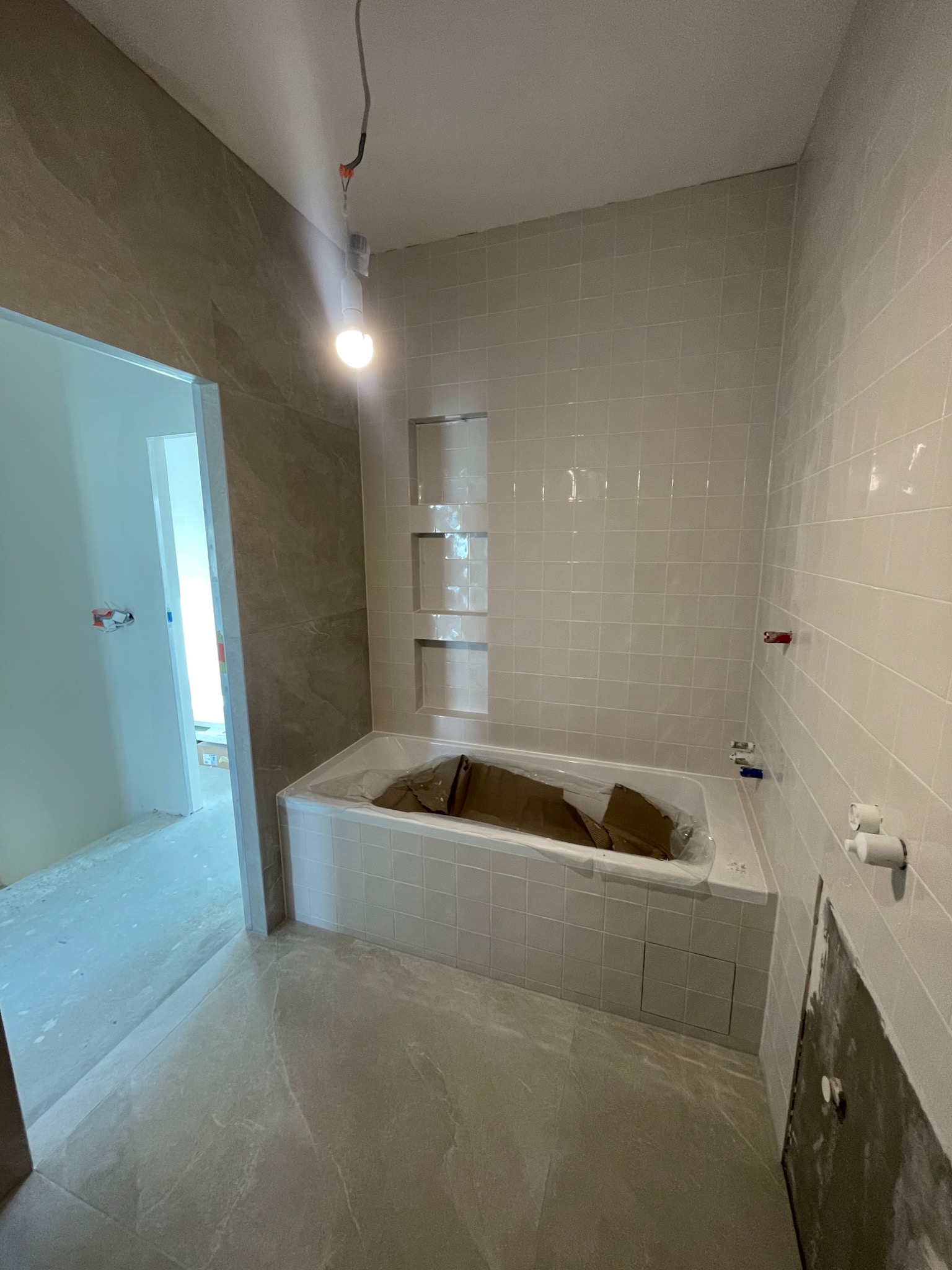 Łazienka w trakcie remontu: biała wanna zabezpieczona folią, jasne płytki na ścianach i podłodze, widoczne niedokończone prace przy instalacji wodno-kanalizacyjnej oraz oświetlenie budowlane.