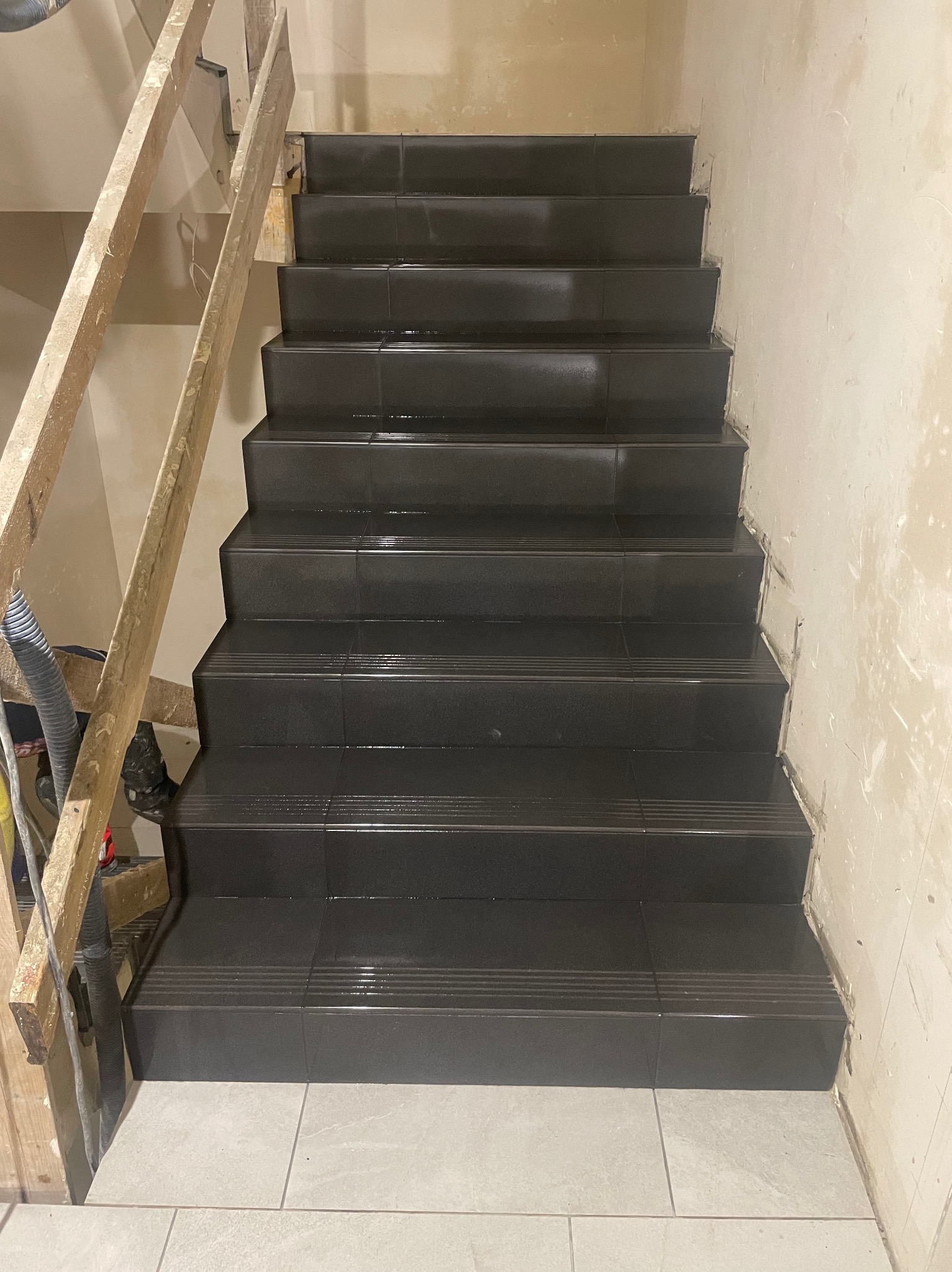 Wykończone schody z ciemnych płytek z ryflowanymi krawędziami, widoczne niedokończone ściany i drewniana konstrukcja pomocnicza.