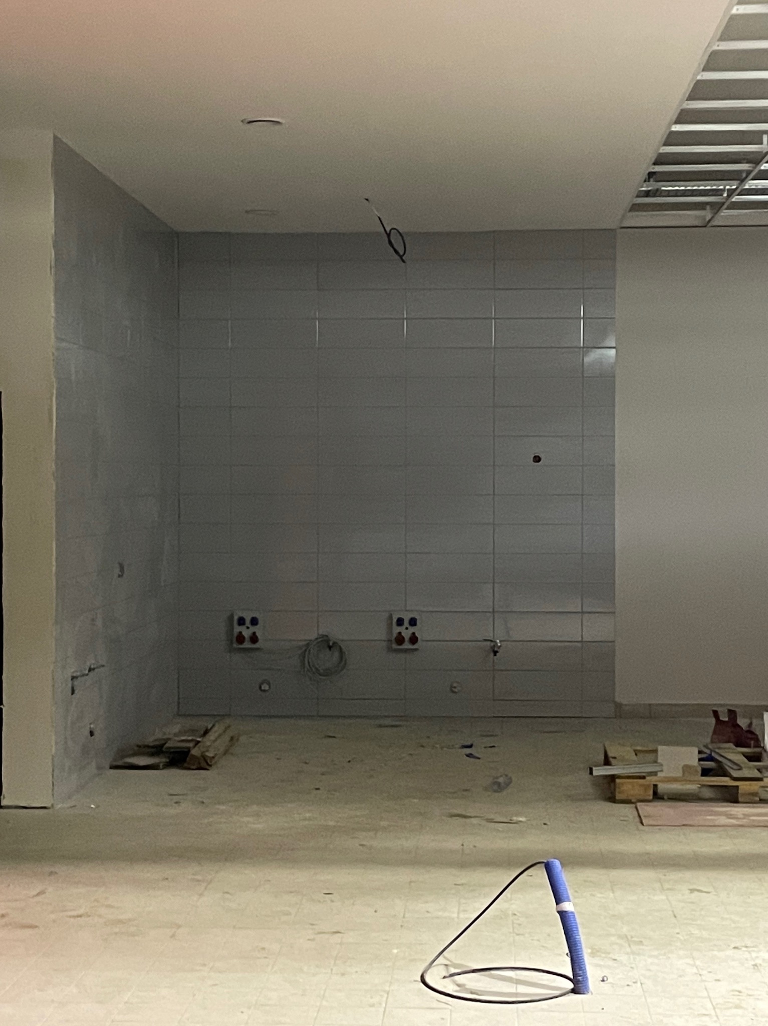 Wnętrze łazienki w trakcie prac wykończeniowych: szare płytki na ścianie, widoczne instalacje, podłoga przygotowana do dalszych prac, narzędzie do cięcia rur na pierwszym planie.