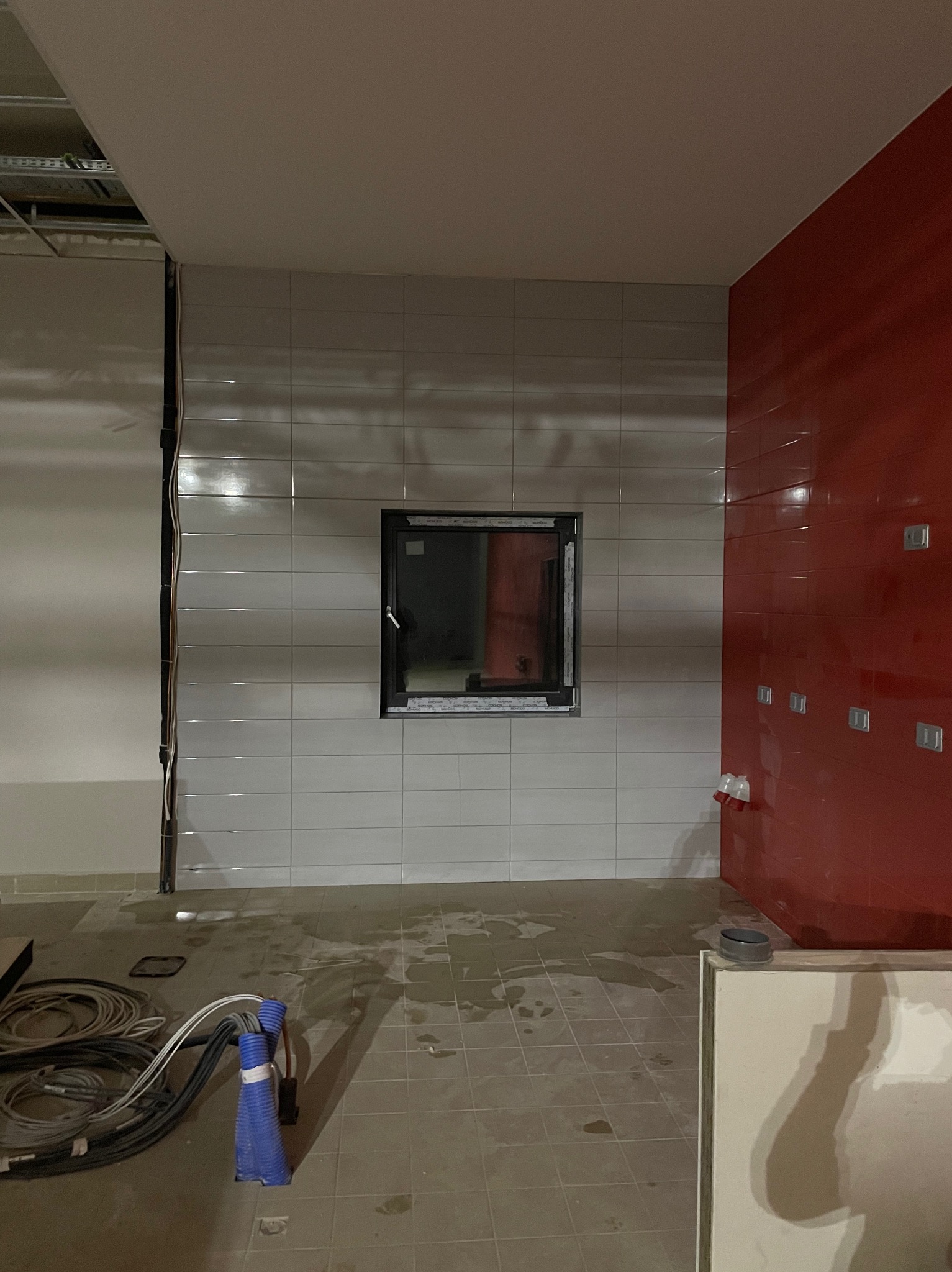 Łazienka w trakcie wykańczania: ułożone jasne płytki na ścianie z oknem, fragment czerwonej ściany z instalacją elektryczną, widoczne kable i narzędzia na podłodze.