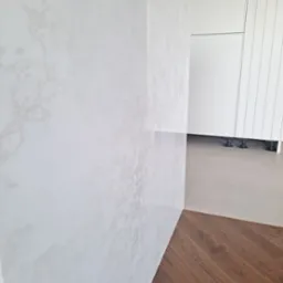 Fragment blatu kuchennego z białego konglomeratu Altamonte o delikatnym, marmurkowym wzorze, przylegający do podłogi z parkietu ułożonego we wzór jodełki, z widocznymi białymi szafkami kuchennymi...
