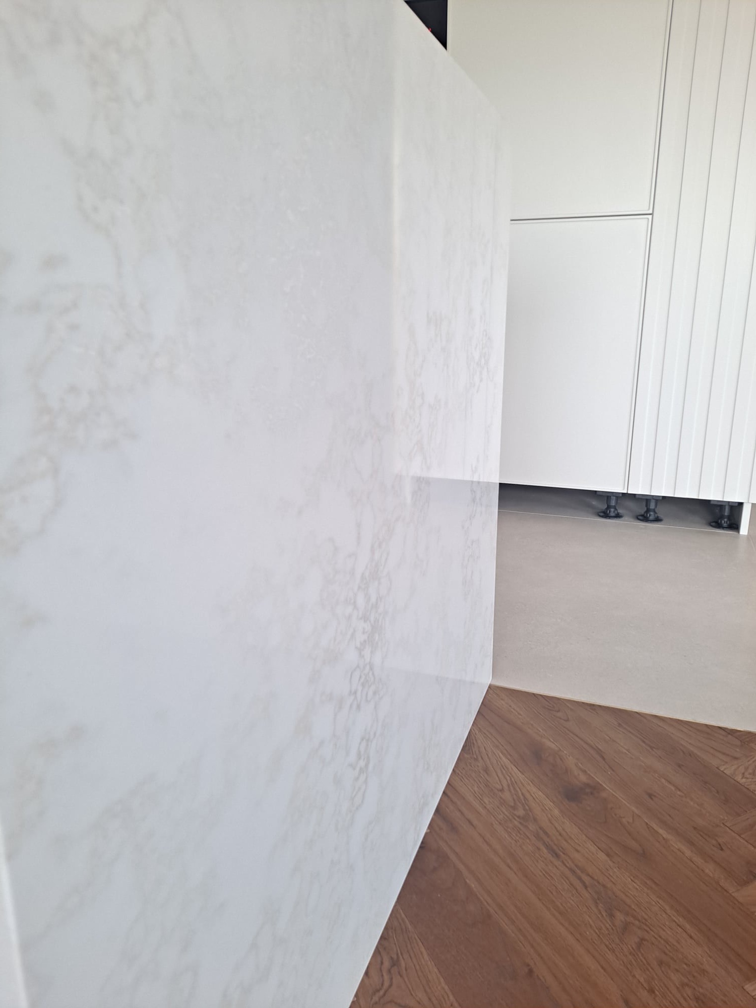 Fragment blatu kuchennego z białego konglomeratu Altamonte o delikatnym, marmurkowym wzorze, przylegający do podłogi z parkietu ułożonego we wzór jodełki, z widocznymi białymi szafkami kuchennymi...